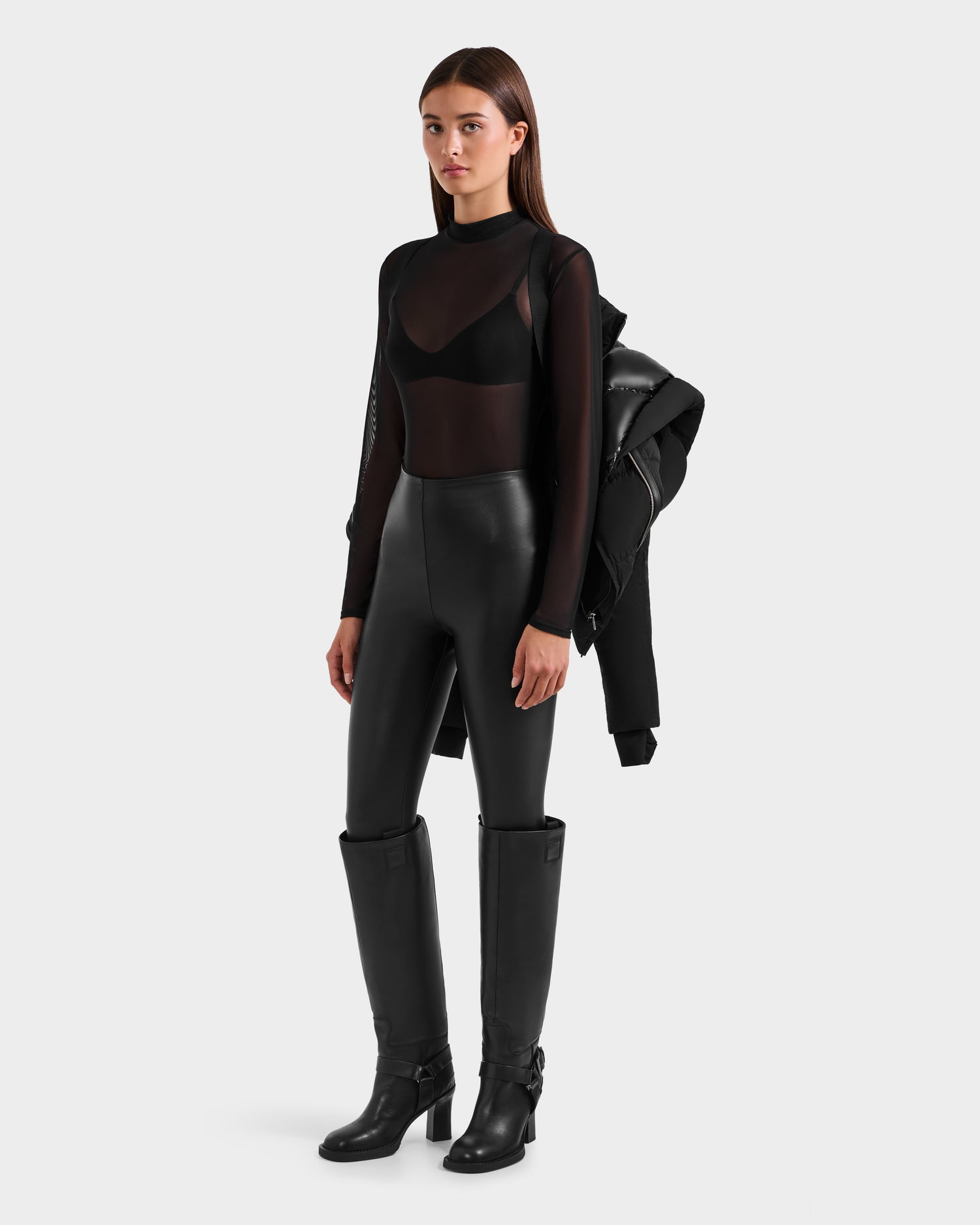 A woman poses against a plain background wearing the Rudsak SHALI - 8124588 Black RDS-certified down short puffer over one shoulder, styled with a sheer black long-sleeve top, black bra, high-waisted pants, and knee-high boots for a fitted look.
