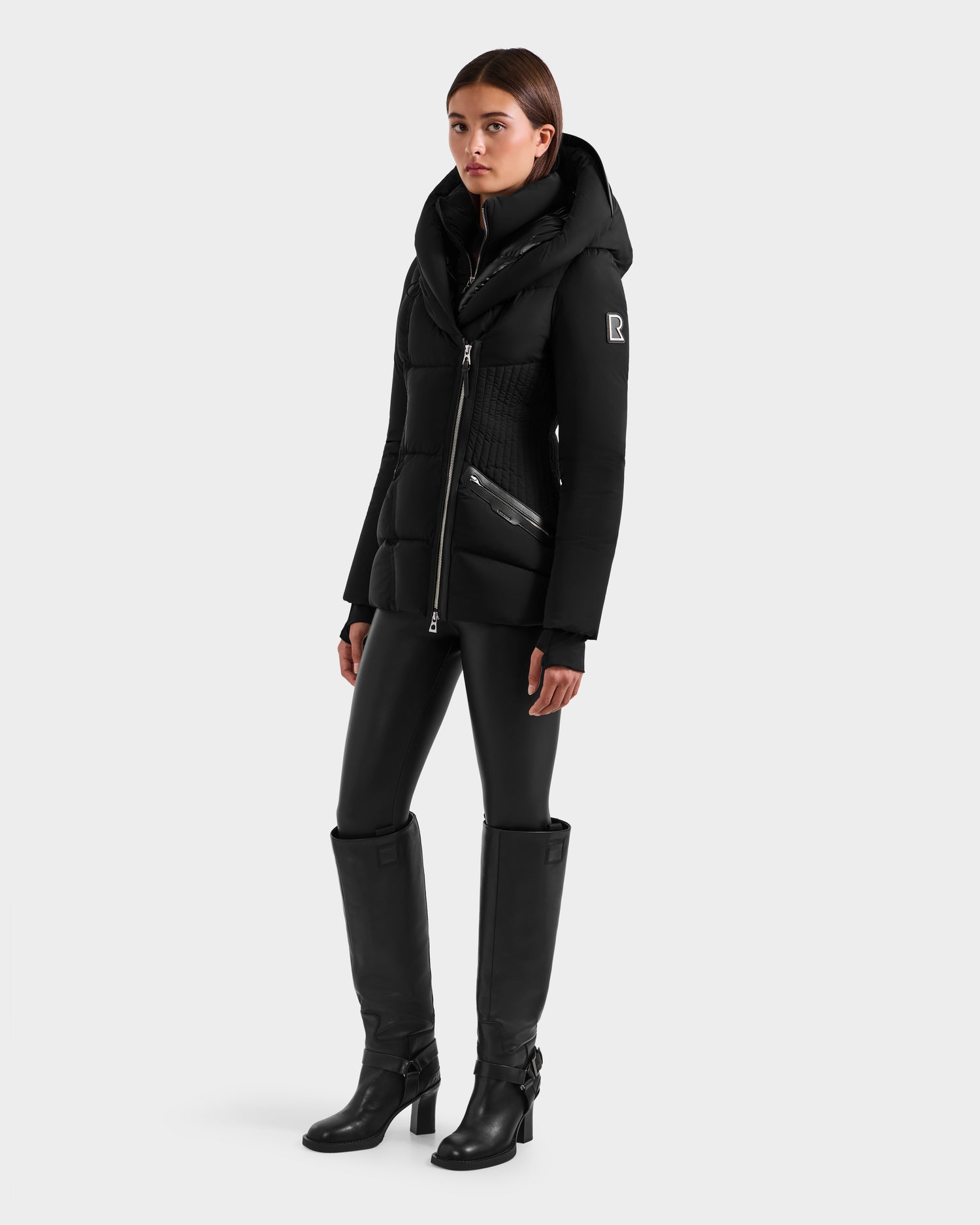 A woman faces forward against a light gray background, wearing the Rudsak SHALI - 8124588 Black women's down puffer jacket with a fitted silhouette, paired with black fitted pants and knee-high black heeled boots.