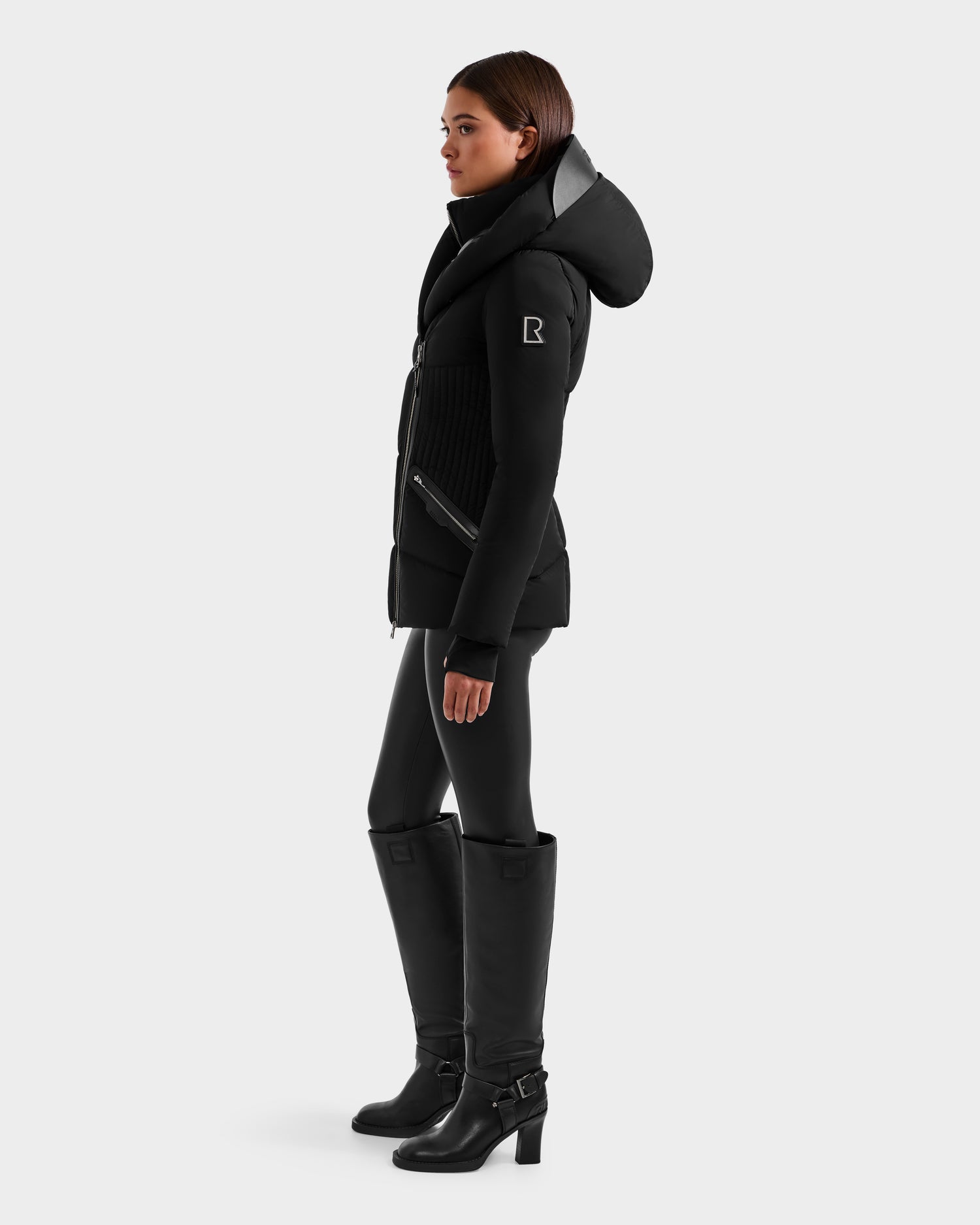A woman stands in profile against a plain light gray background, wearing the Rudsak SHALI - 8124588 Black women's down puffer jacket with a high collar, styled with black leggings and tall black heeled boots for a sleek, fitted silhouette.