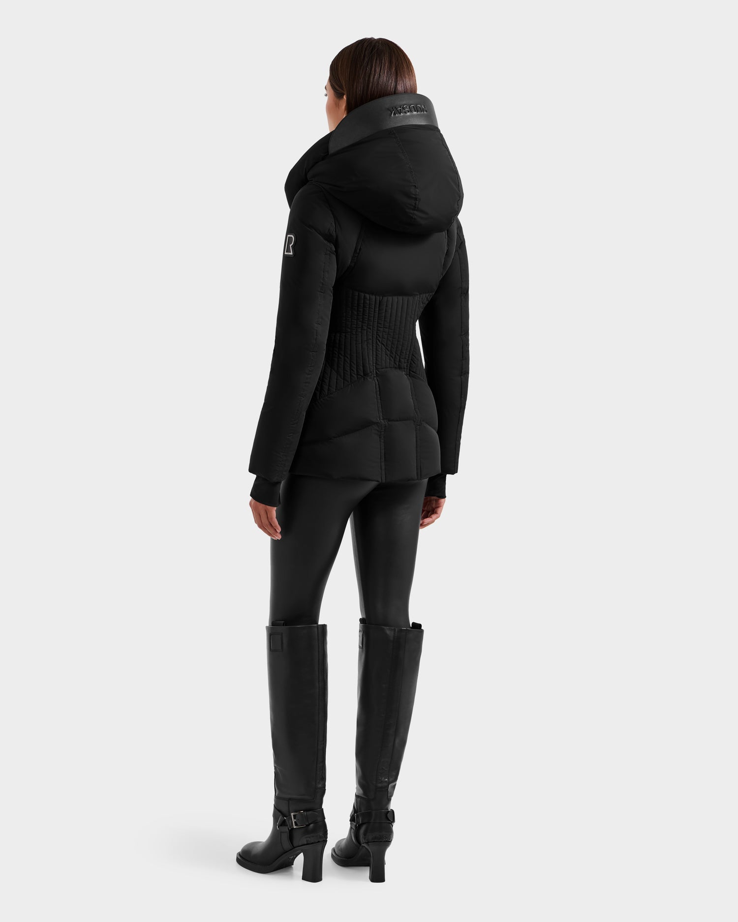 A person models the Rudsak SHALI - 8124588 Black women's down puffer jacket with a fitted silhouette, paired with black leggings and tall boots, standing against a plain light background.