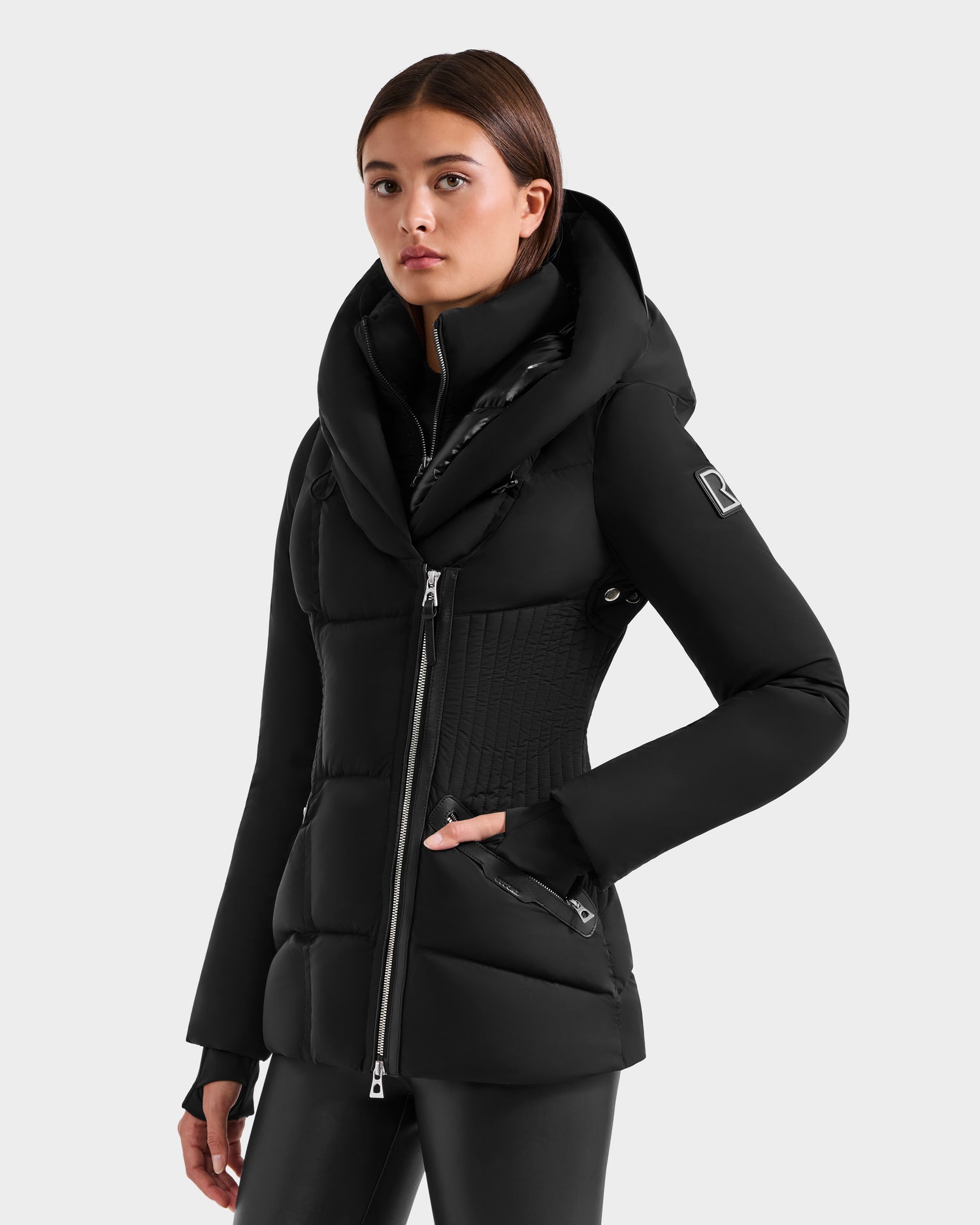 A young woman wears the Rudsak SHALI - 8124588 Black women’s down puffer jacket with a zip-up front, large hood, and fitted silhouette, posing with one hand in her pocket against a plain light gray background.