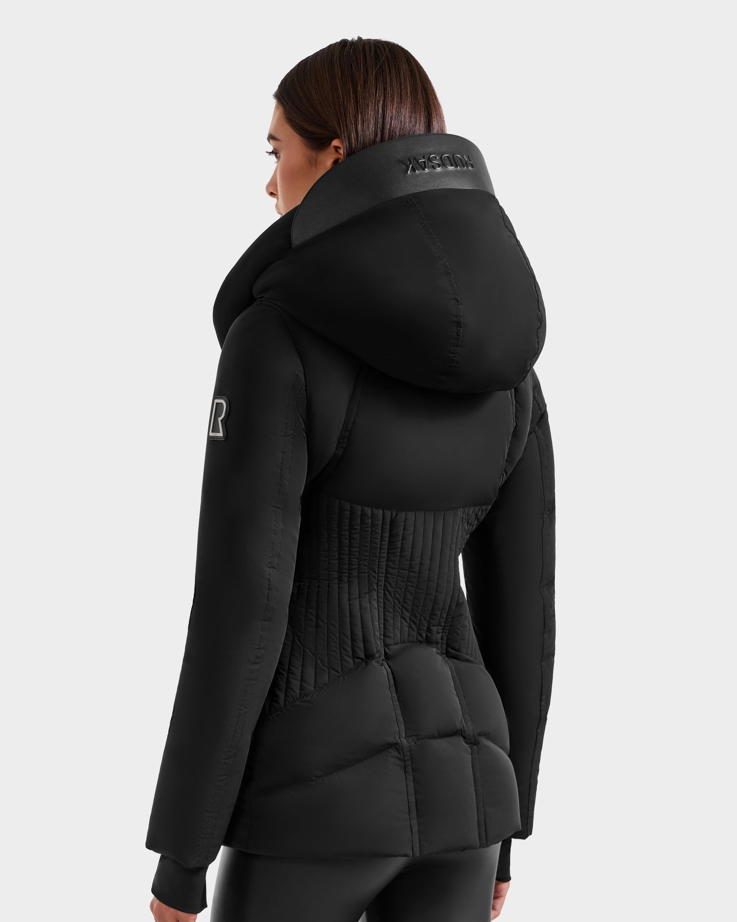 A woman with straight brown hair models the Rudsak SHALI - 8124588 Black fitted winter jacket, featuring a high collar, hood, and quilted details. Brand logos are visible on the upper back and sleeve against a plain light gray background.