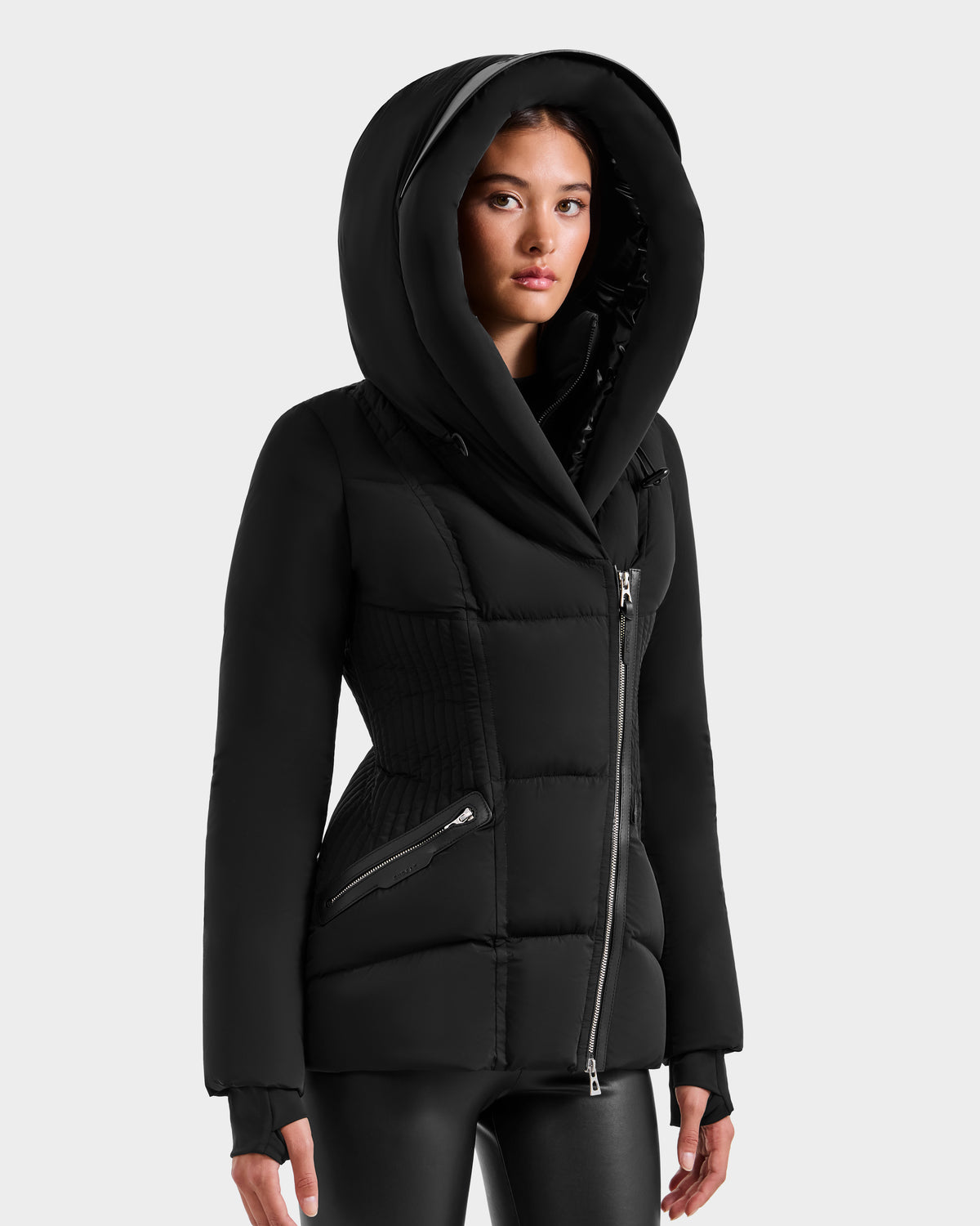 A woman models the Rudsak SHALI - 8124588 Black puffer jacket, featuring a fitted silhouette, large hood, and asymmetrical zipper. She stands against a light gray background with straight dark hair and a neutral expression.