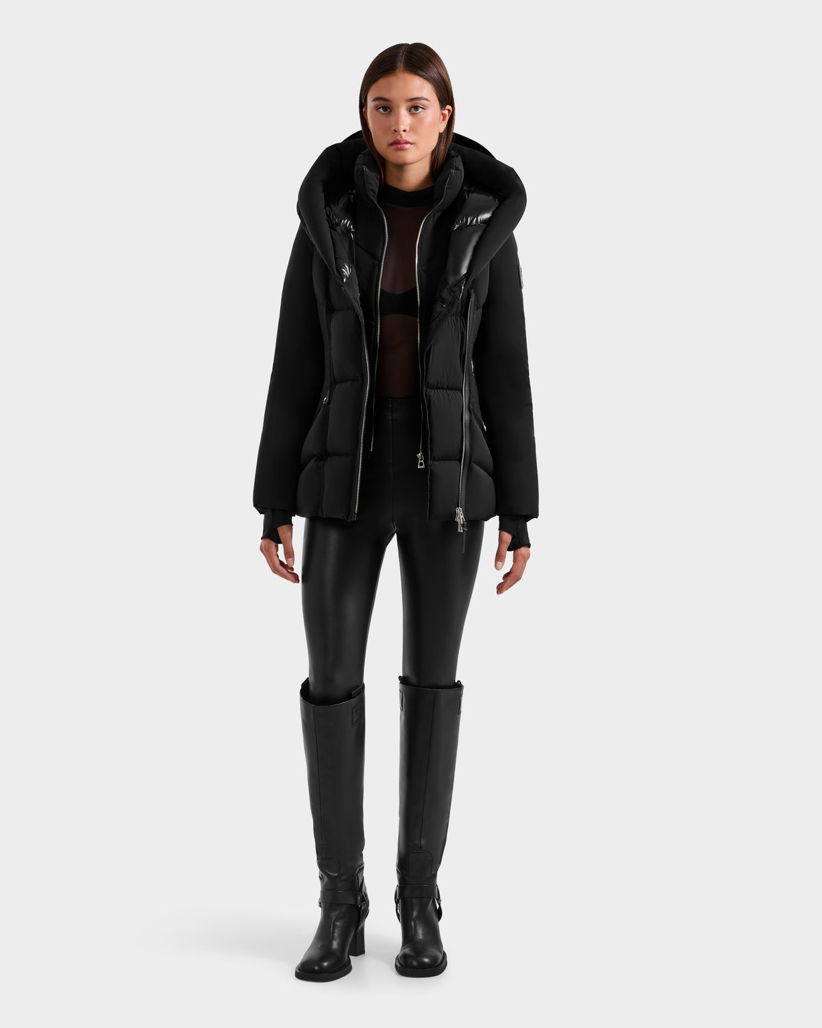A woman with straight brown hair and a neutral expression stands against a plain background, wearing the Rudsak SHALI - 8124588 Black women's short down puffer, a black sheer top, black leather pants, and knee-high boots.