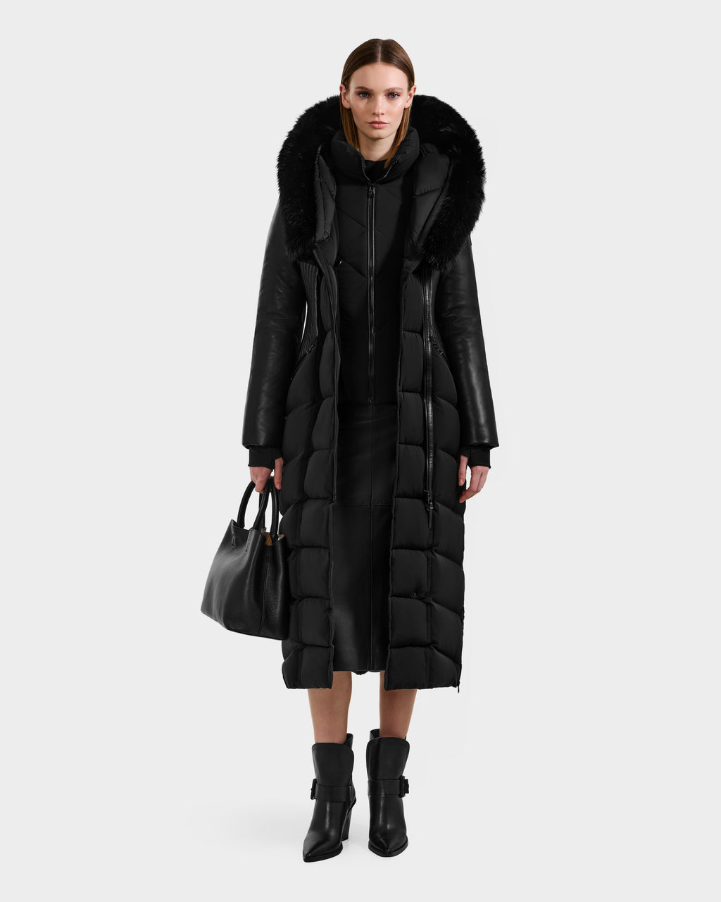 A woman faces forward in a Rudsak SHANNON - 8121511 BLACK fitted puffer coat with a fur-trimmed hood, styled over a black dress and ankle boots while carrying a black handbag against a plain light gray background.