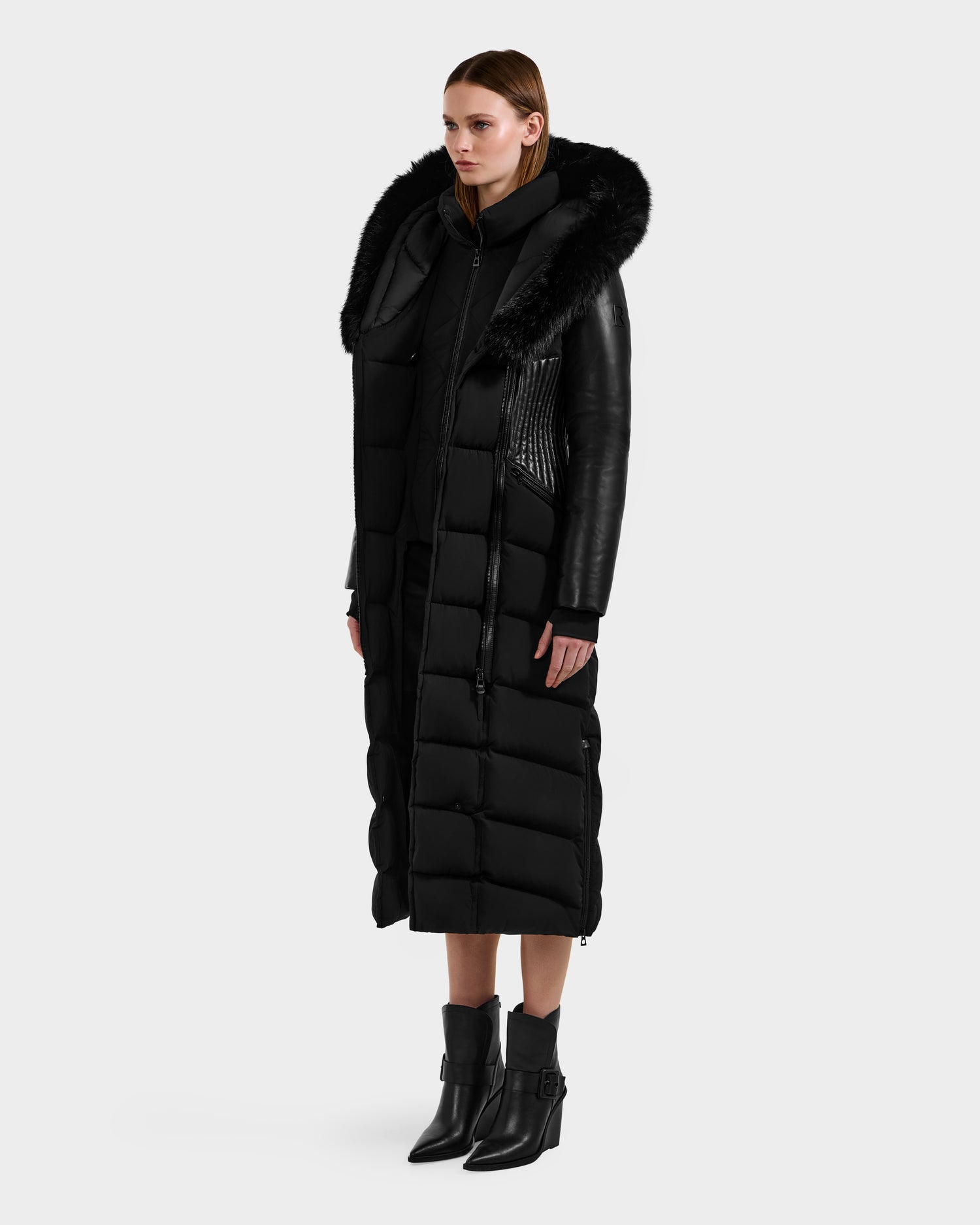 A woman faces forward against a plain light background, wearing the Rudsak SHANNON - 8121511 BLACK quilted maxi down parka with leather sleeves and fur-trimmed hood, paired with black heeled ankle boots featuring buckles.