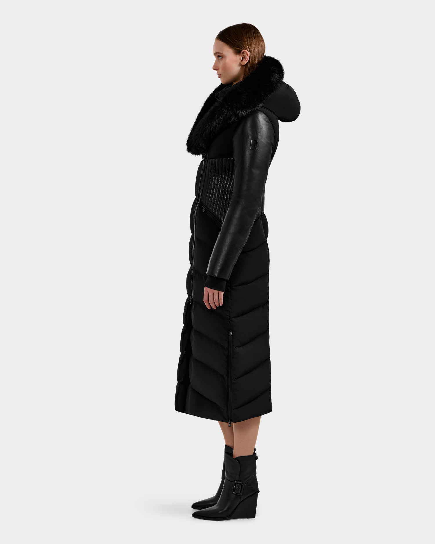 A woman stands in profile wearing the Rudsak SHANNON - 8121511 BLACK, a fitted puffer with leather sleeves, a large fur collar, and heeled ankle boots with buckles. The knee-length coat is a chic winter essential.