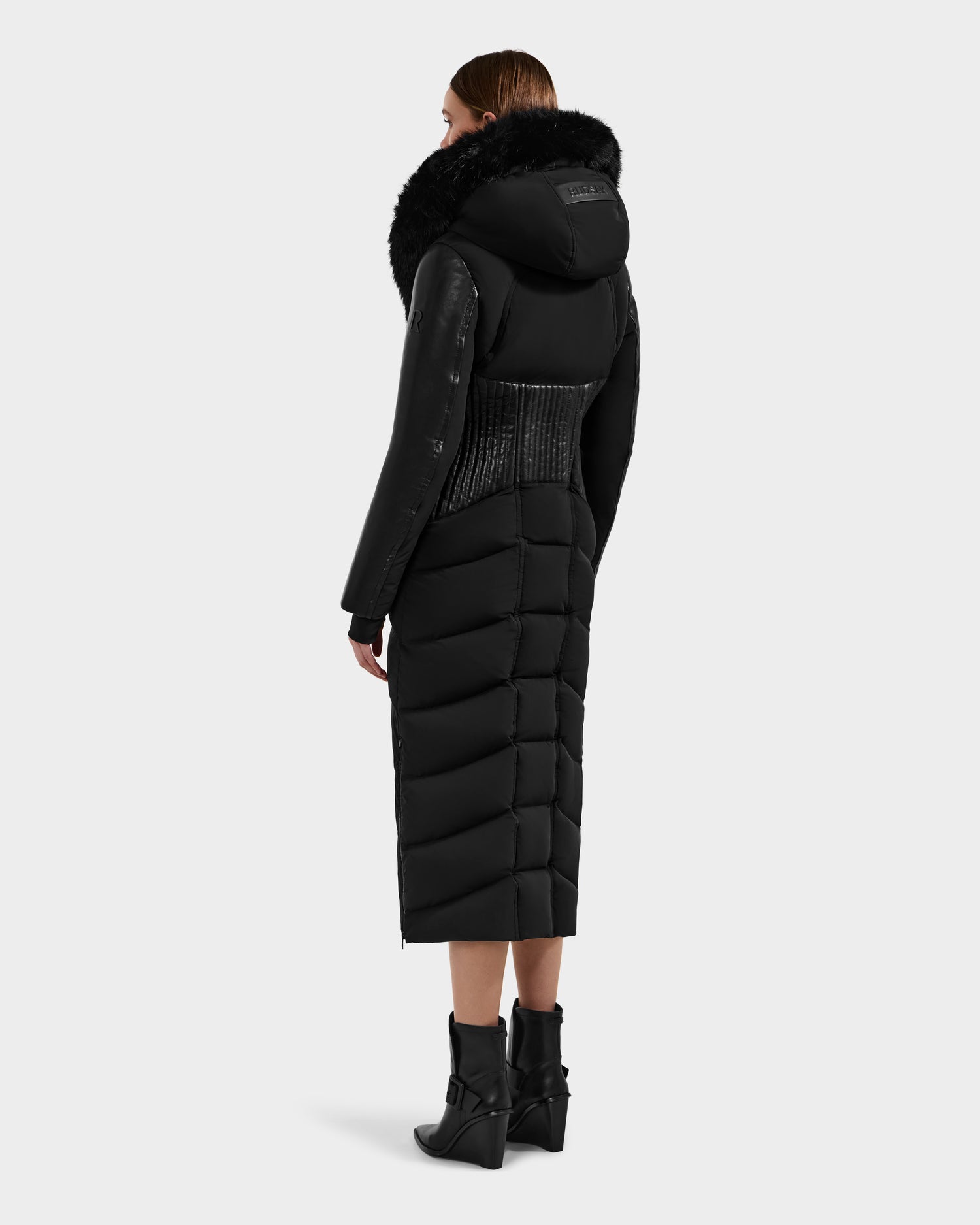 A woman faces away, wearing the Rudsak SHANNON - 8121511 BLACK fitted puffer coat with a textured waist and large fur-trimmed hood, paired with black ankle boots, set against a plain light gray background.