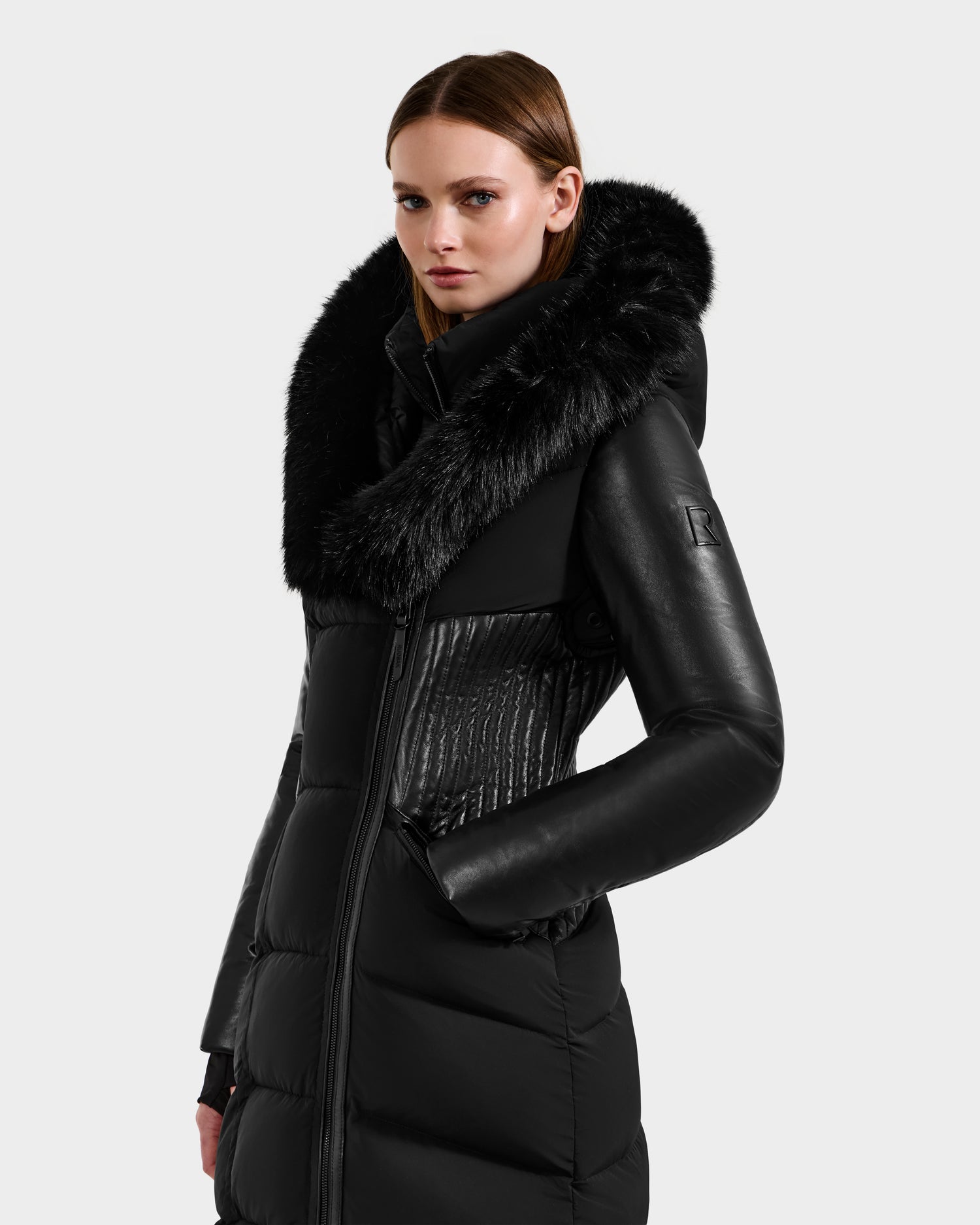 A woman wears the Rudsak SHANNON - 8121511 BLACK, a stylish black quilted maxi down parka with leather sleeves and a large faux fur collar, posing against a plain light gray background.