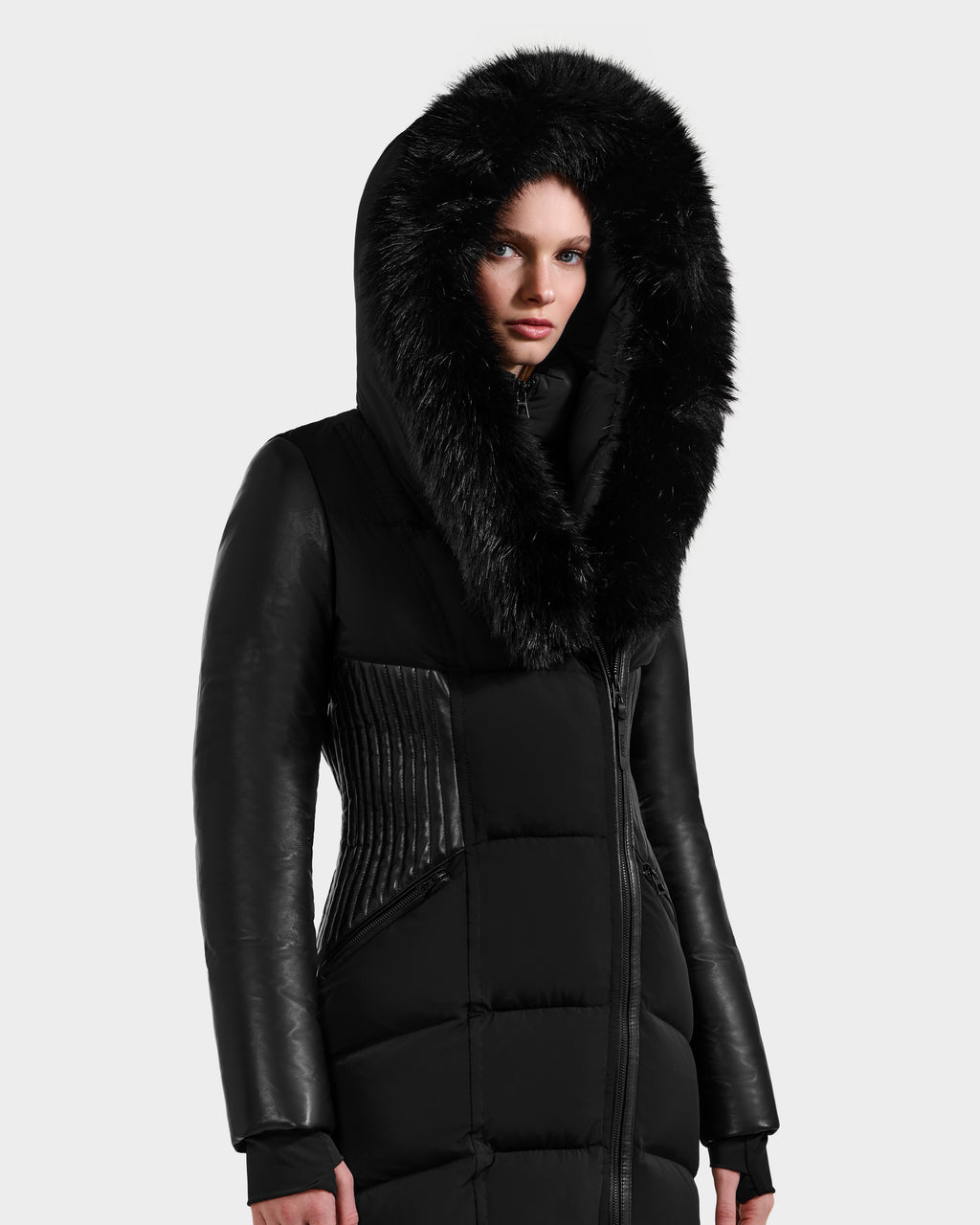 A woman wears the Rudsak SHANNON - 8121511 BLACK fitted puffer coat with a large fur-lined hood, quilted padding, and leather-textured sleeves as she faces forward against a plain light gray background.