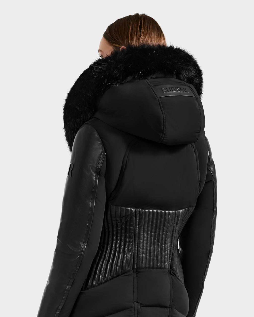 A person is seen from behind in a Rudsak SHANNON - 8121511 BLACK jacket, featuring a large fur-lined hood, ribbed back detailing, and sleek leather-like sleeves, set against a plain light background.