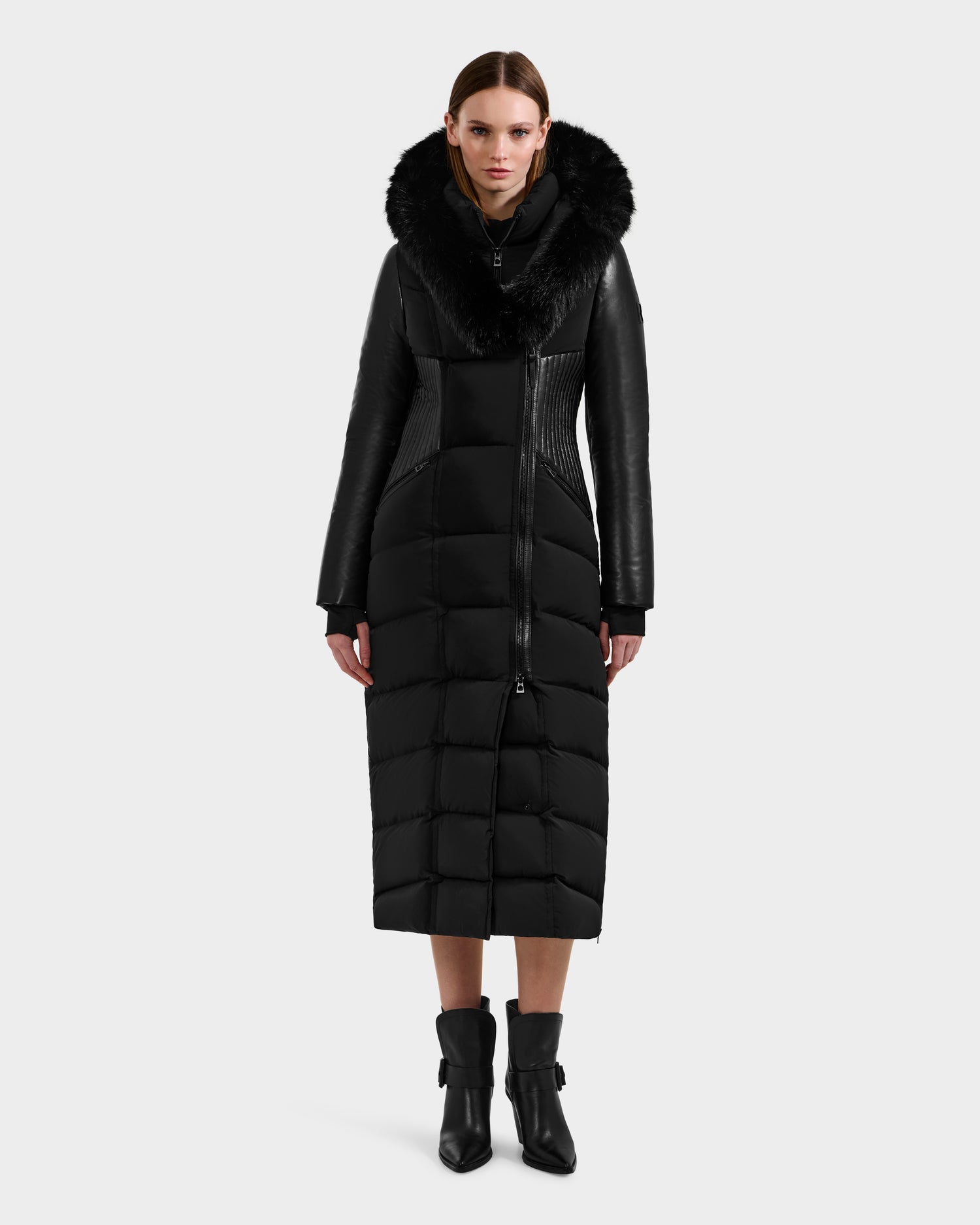 A woman faces forward against a light background, wearing the Rudsak SHANNON - 8121511 BLACK fitted puffer coat with a large black fur-trimmed hood and dark ankle boots, her hands resting by her sides.
