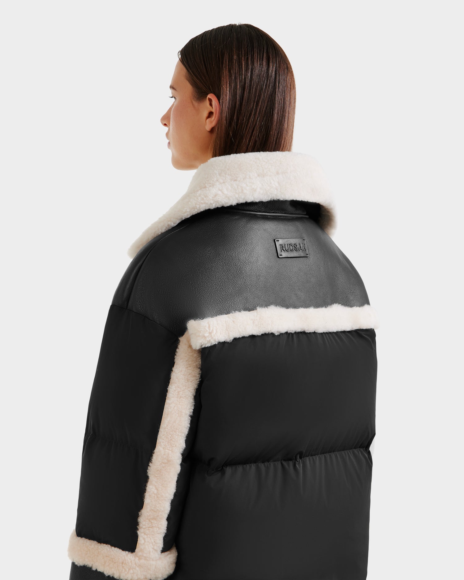 A person with straight brown hair is seen from behind wearing the Rudsak SHIRLEY - 8125613 Black womens down jacket, featuring white faux shearling trim and collar, with a rectangular patch on the upper back.