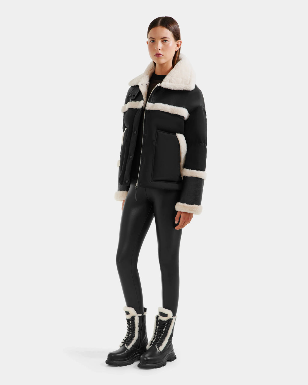 A woman with straight dark brown hair and a neutral expression wears the Rudsak SHIRLEY - 8125613 Black shearling jacket with white trim, black leggings, and black lace-up boots with white shearling accents against a plain background.