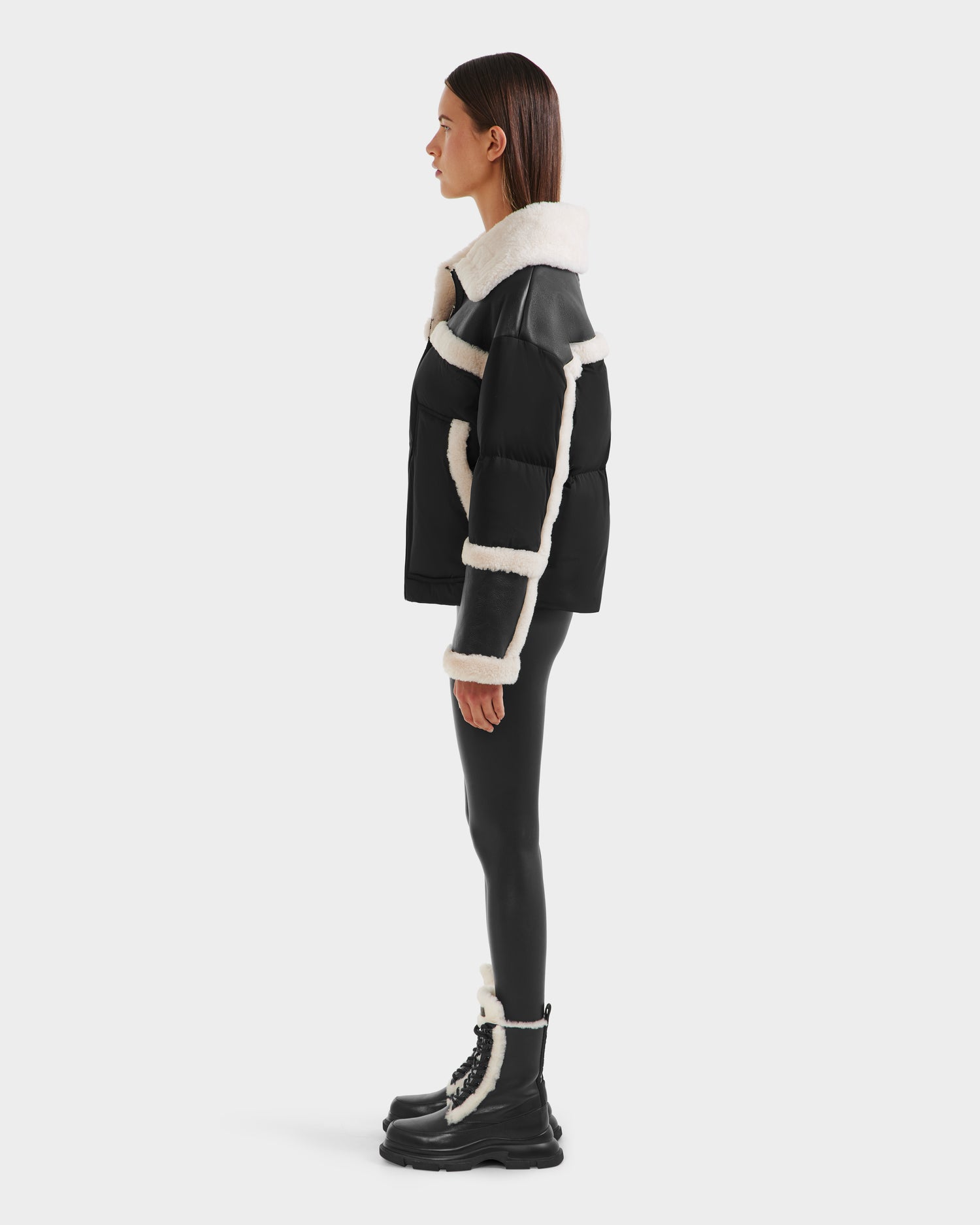 A person in profile showcases chic winter style wearing Rudsak’s SHIRLEY - 8125613 Black shearling jacket, glossy black leggings, and black lace-up boots with white trim, all set against a plain white background.