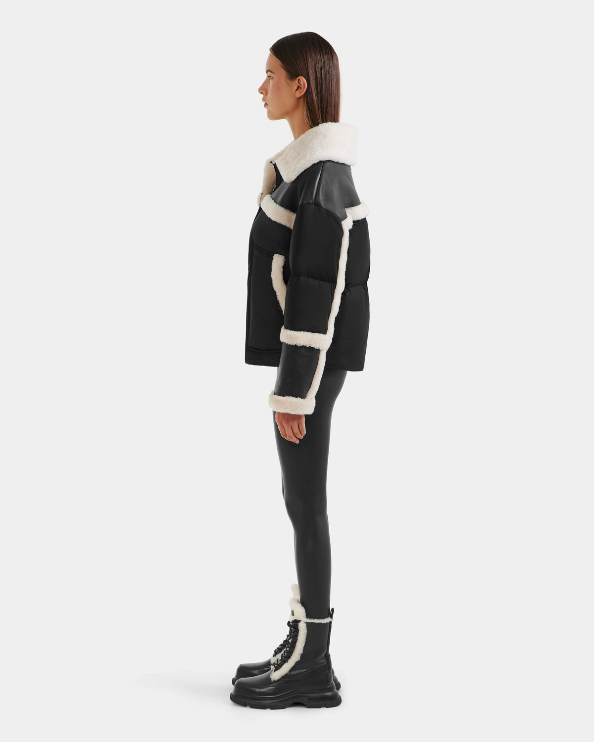 A person in profile showcases chic winter style wearing Rudsak’s SHIRLEY - 8125613 Black shearling jacket, glossy black leggings, and black lace-up boots with white trim, all set against a plain white background.