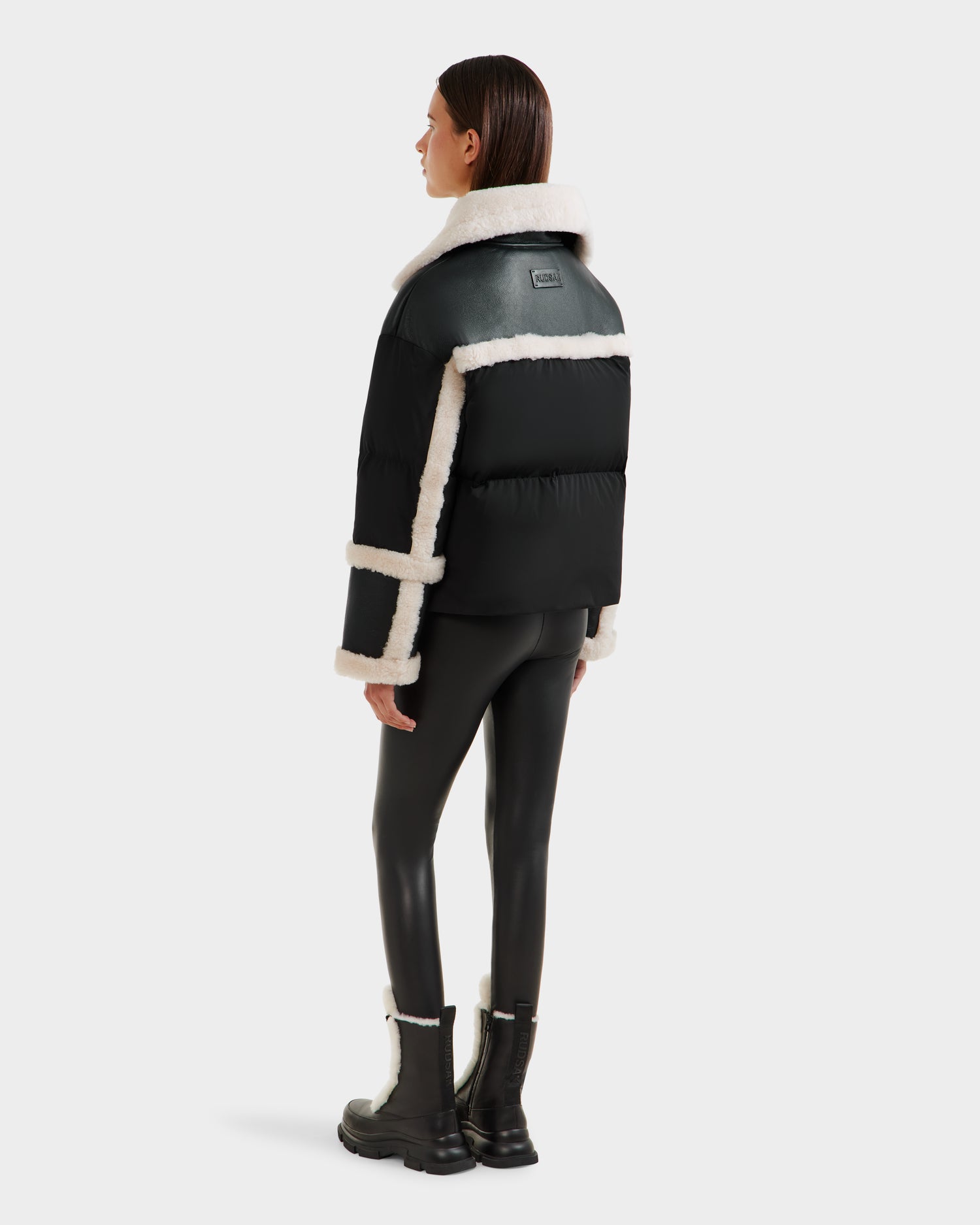 A woman, seen from behind, wears the Rudsak SHIRLEY - 8125613 Black cropped winter jacket with white shearling trim, black leggings, and black boots with white shearling details against a plain white background.