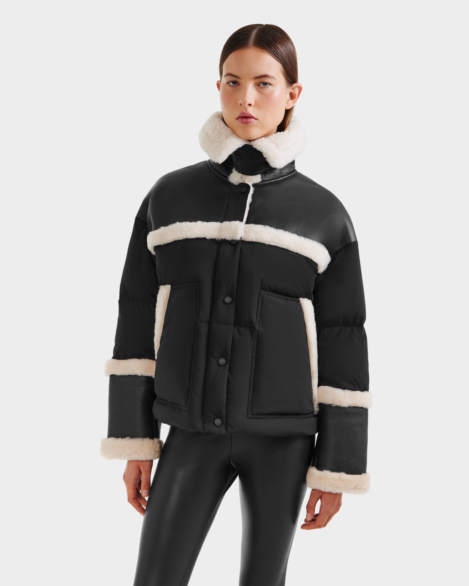 A woman models the Rudsak SHIRLEY - 8125613 Black women’s down jacket with white shearling trim, paired with black leather pants against a plain white background—perfect for winter style.