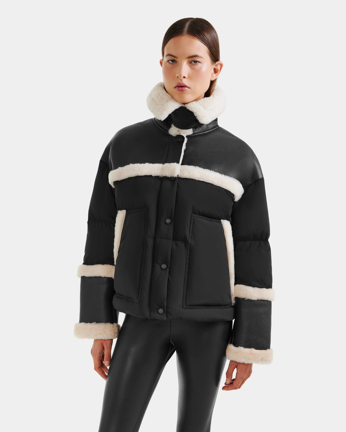 A woman models the Rudsak SHIRLEY - 8125613 Black women’s down jacket with white shearling trim, paired with black leather pants against a plain white background—perfect for winter style.