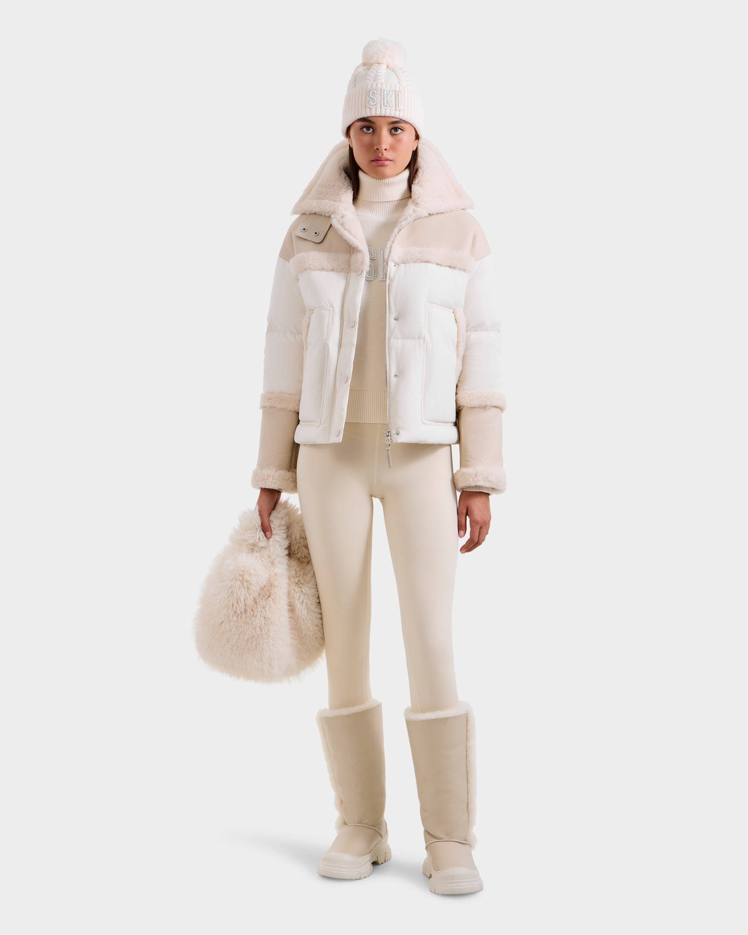 A person wearing the Rudsak SHIRLEY - 8125613 Vanilla cropped winter jacket, paired with a knit hat, leggings, fur boots, and holding a fluffy bag stands against a plain white background.