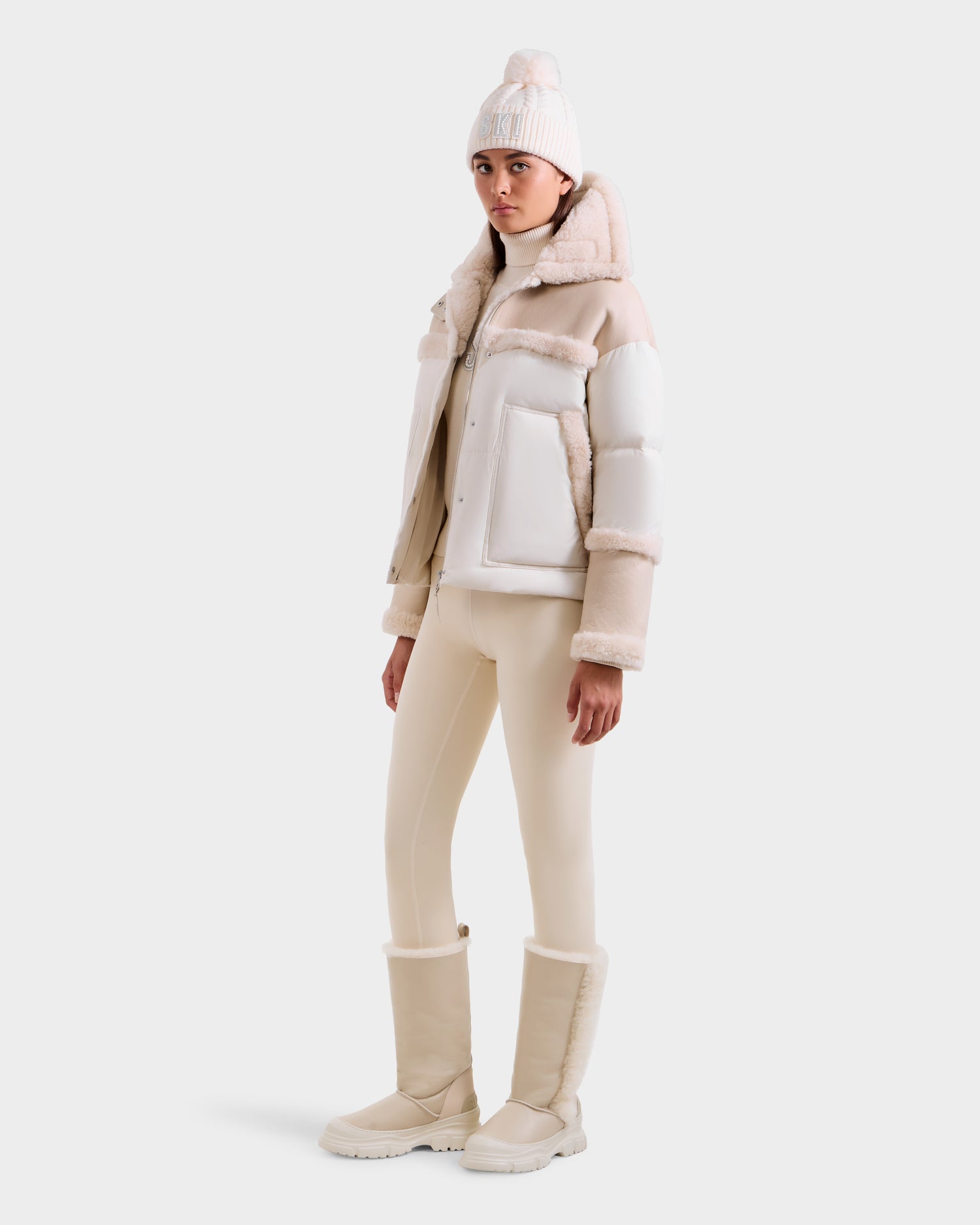 A person stands slightly turned against a plain background wearing the SHIRLEY - 8125613 Vanilla boots by Rudsak, paired with a cream beanie with pom-pom, cropped shearling jacket, and cream leggings.
