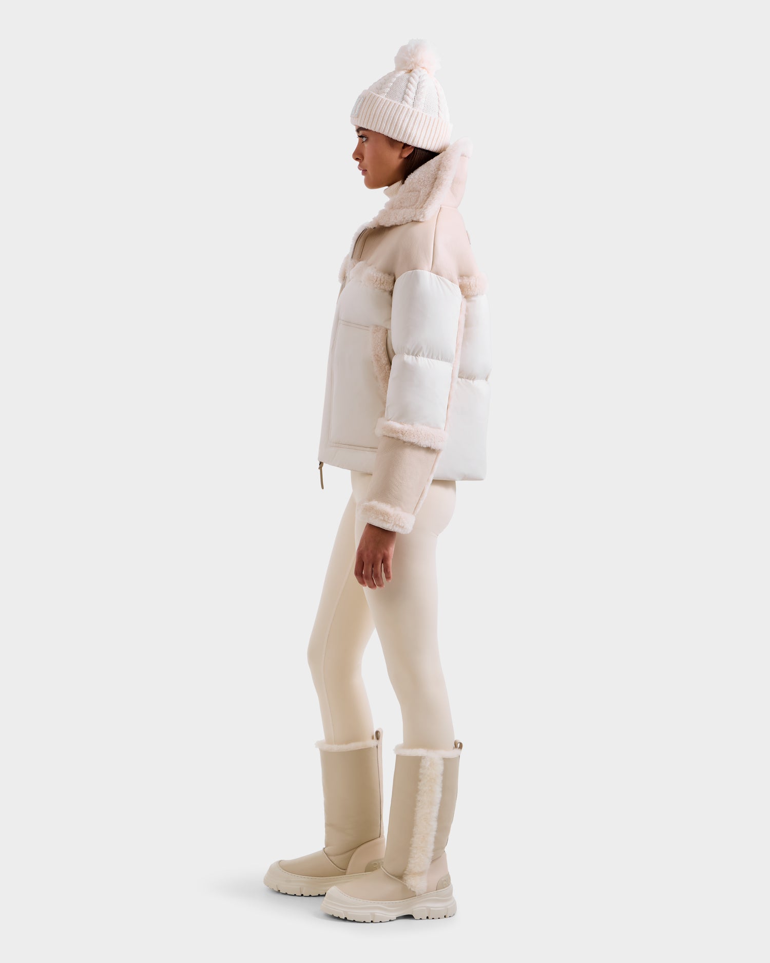 A person stands in profile wearing the Rudsak SHIRLEY - 8125613 Vanilla women’s down jacket with fur trim, cream leggings, matching fur-trimmed boots, and a knit pom-pom beanie against a plain light background.