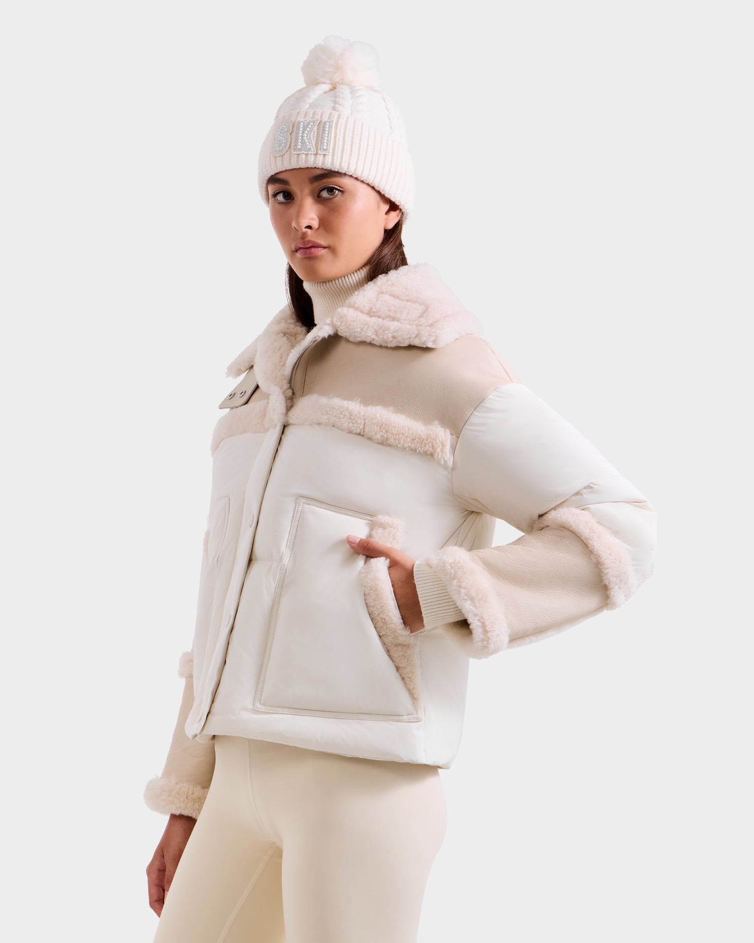 A woman models the Rudsak SHIRLEY - 8125613 Vanilla shearling jacket, paired with a cream pom-pom beanie and light beige leggings, showcasing stylish winterwear against a minimalist backdrop.