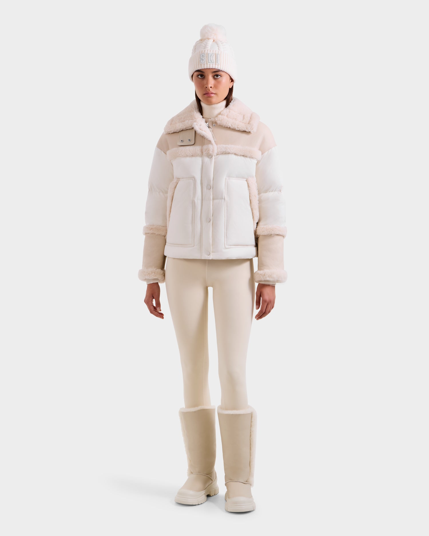 A woman wearing the Rudsak SHIRLEY - 8125613 Vanilla down jacket, a knit hat, cream leggings, and tall winter boots stands against a plain white background.