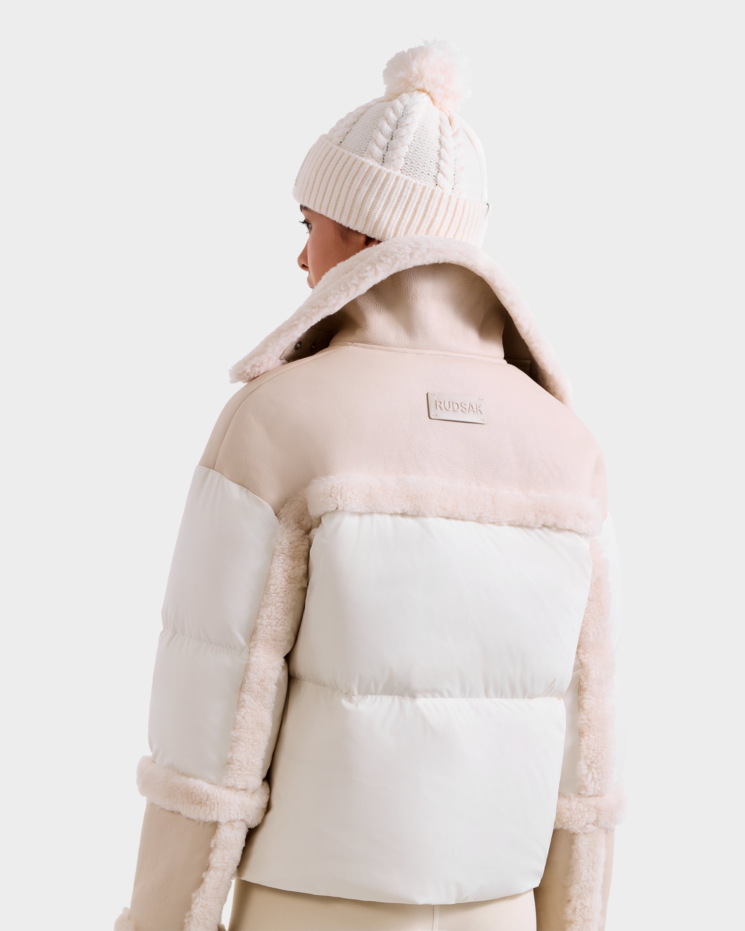 A person is shown from the back wearing the Rudsak SHIRLEY - 8125613 Vanilla cropped winter jacket with faux fur trim and a knit beanie with a pom-pom, set against a plain light background.