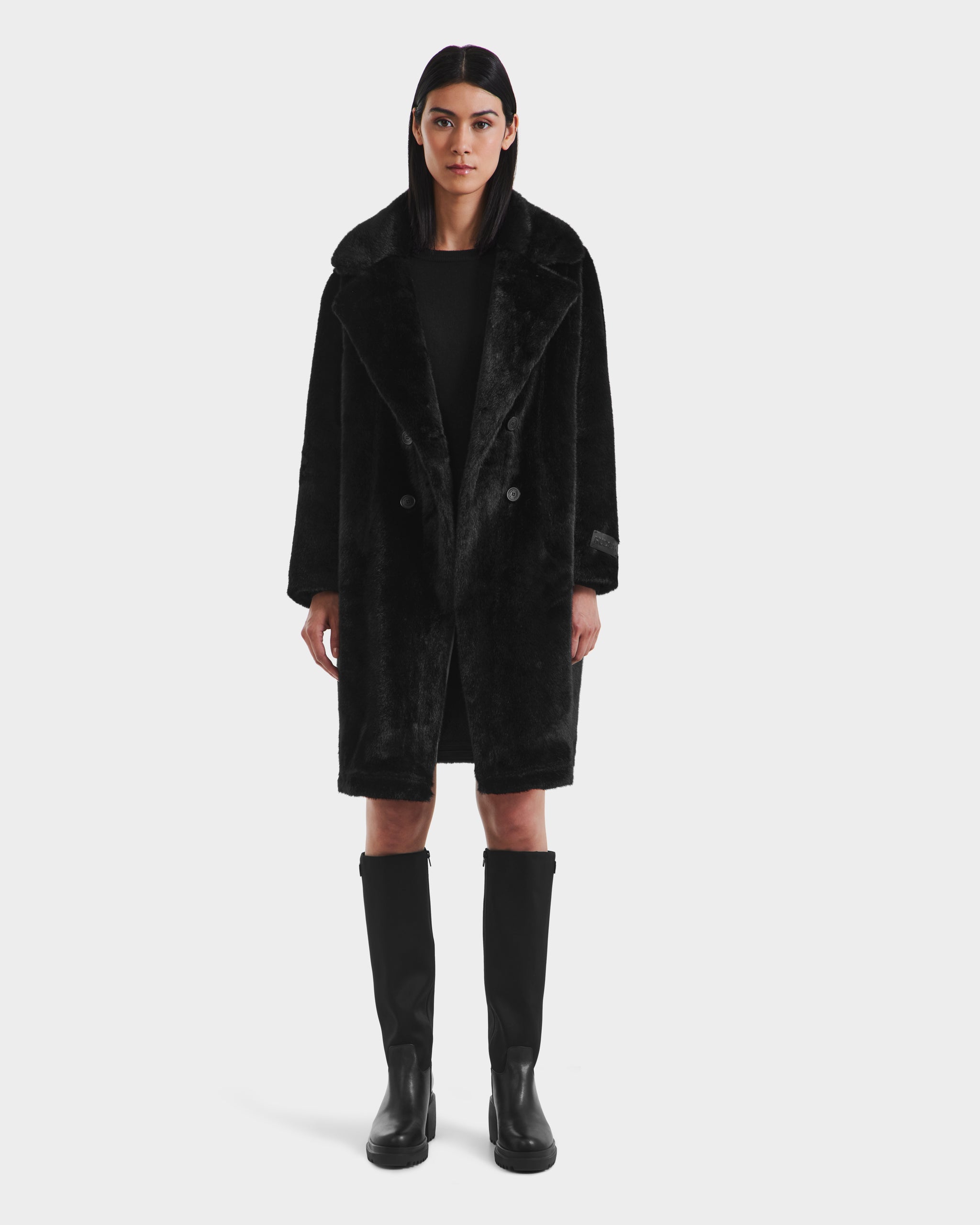 A woman faces forward wearing the Rudsak SIENA - 8125542 Black faux fur coat over a black dress and tall black boots, set against a plain white background.