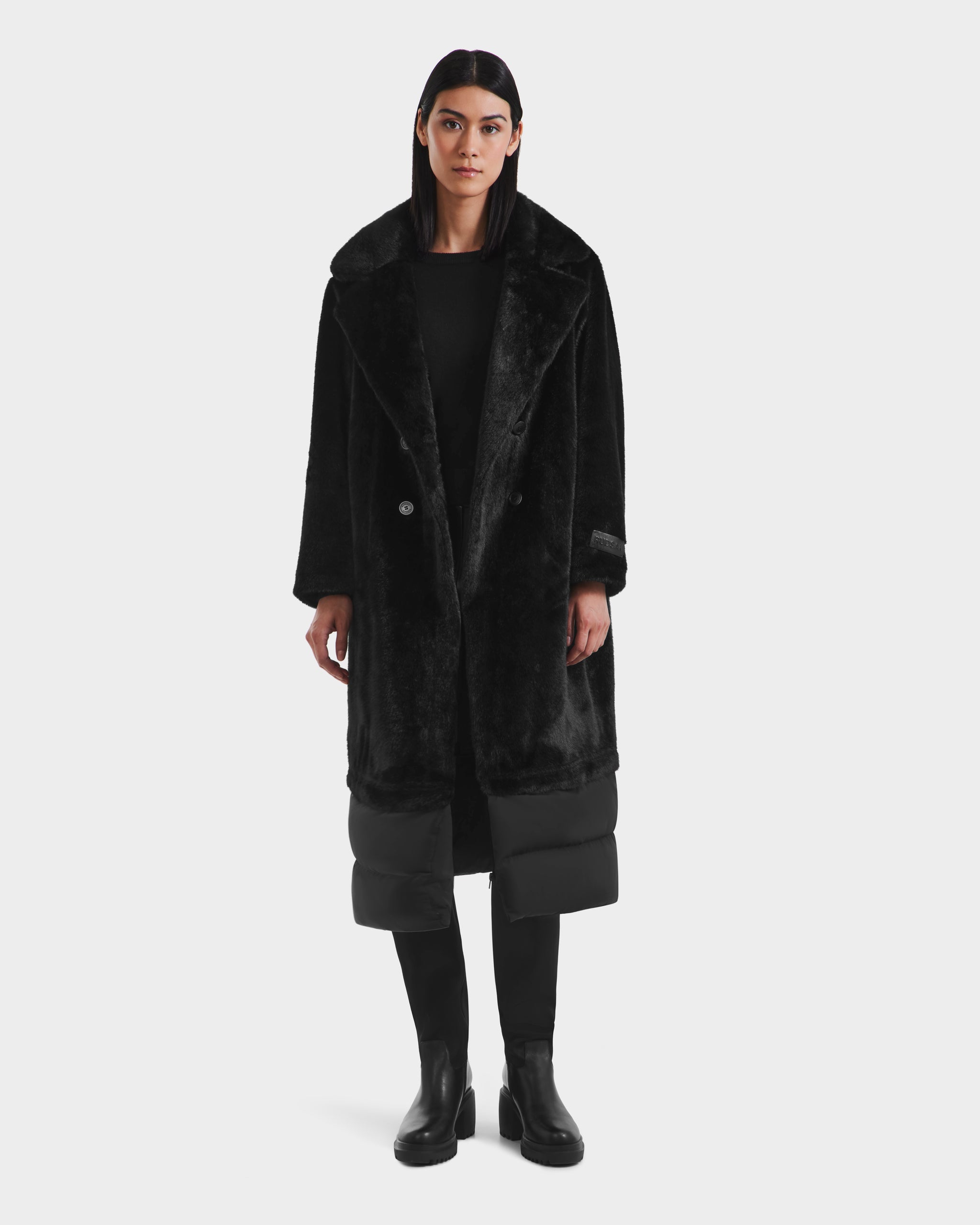A woman with straight dark hair models the Rudsak SIENA - 8125542 Black women’s faux fur winter coat with quilted details, paired with black pants and ankle boots, standing against a plain white background. The coat exudes modern warmth and elegance.