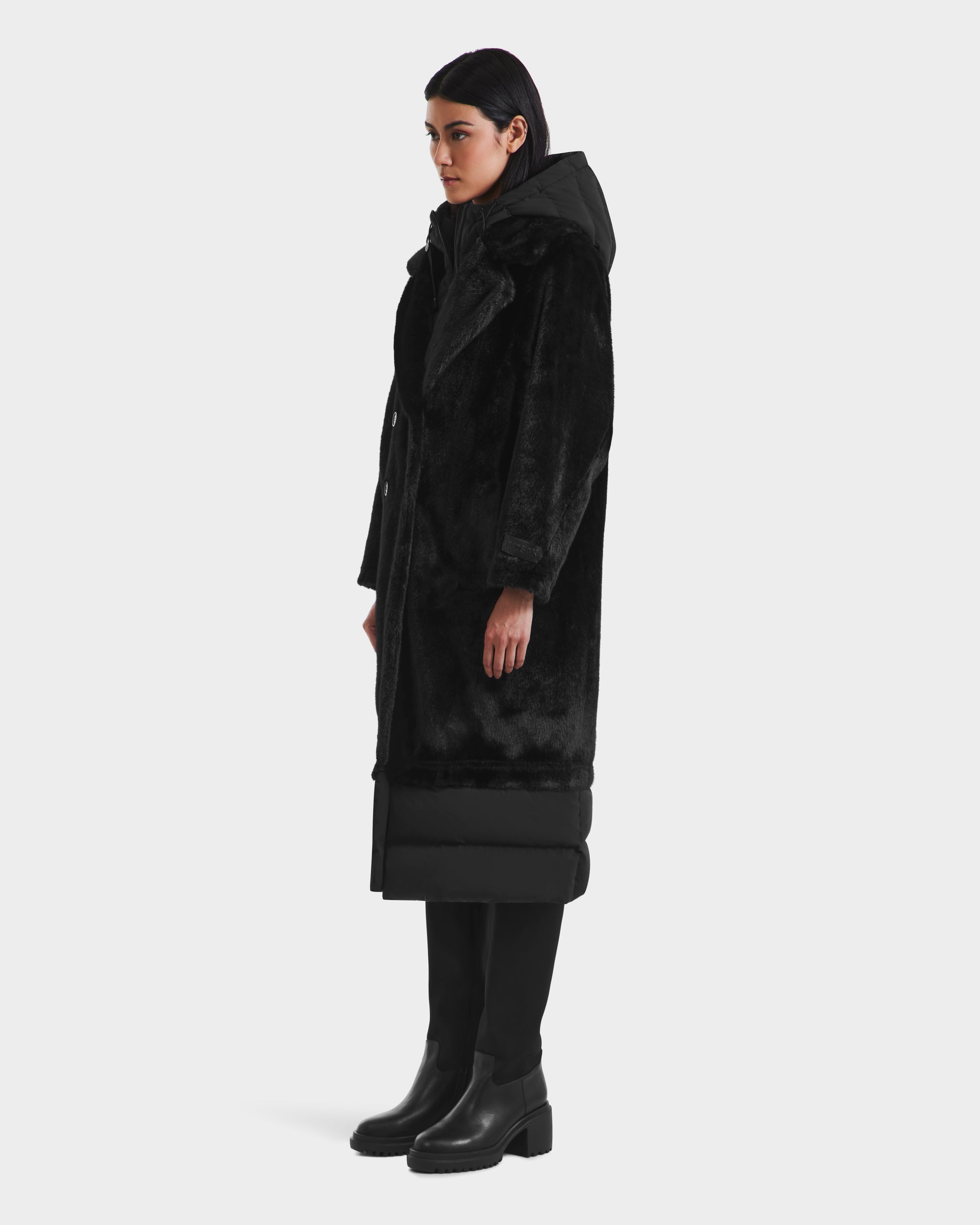 A person with short dark hair stands in profile wearing the Rudsak SIENA - 8125542 Black long hooded winter coat, black pants, and heeled boots against a plain light background.