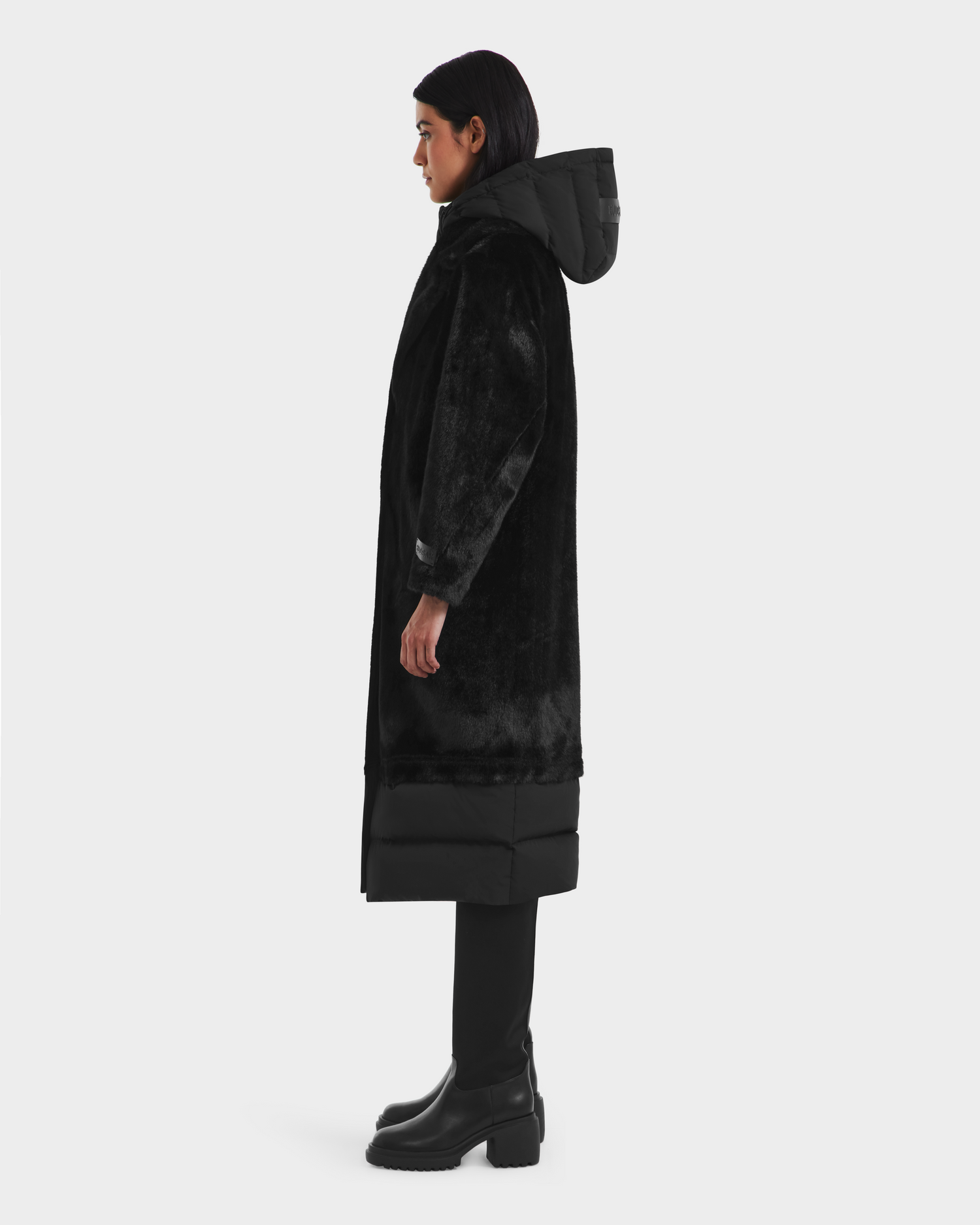 A person stands in profile against a plain light background wearing the Rudsak SIENA - 8125542 Black winter coat with a hood, paired with black pants and boots.