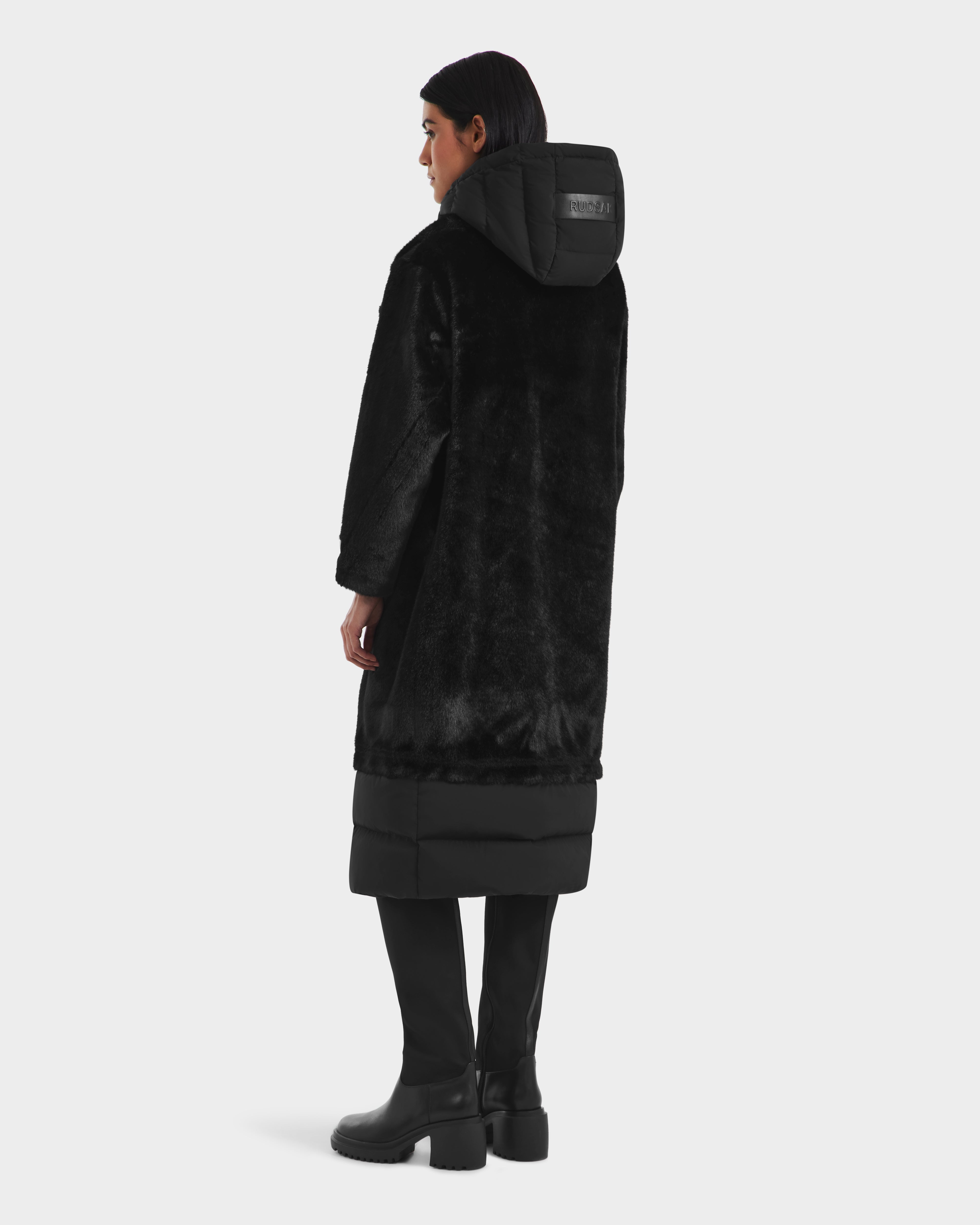 A person stands facing away against a plain white background, wearing the Rudsak SIENA - 8125542 Black women’s faux fur hooded coat, paired with black pants and black heeled boots.