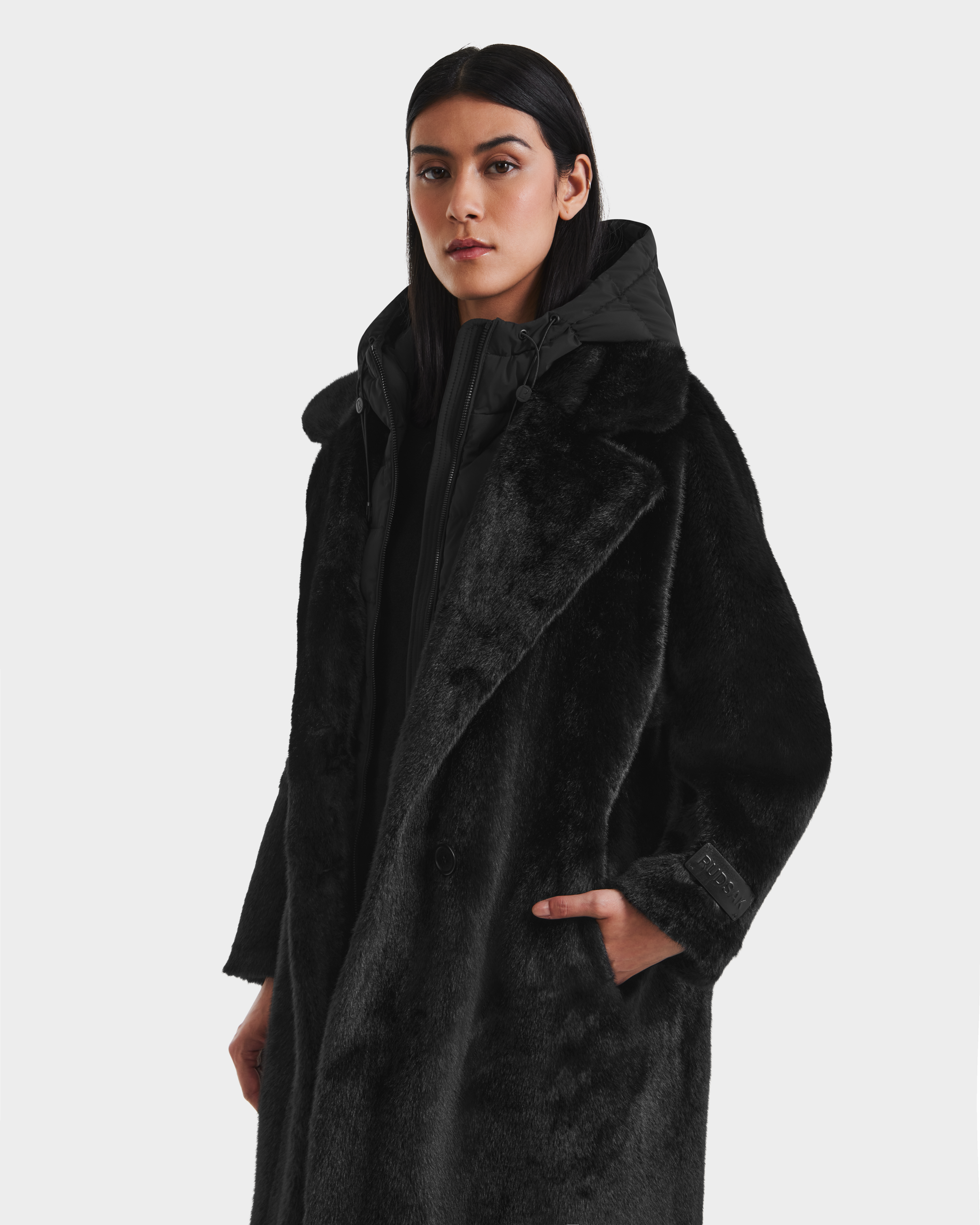 A woman with straight dark hair wears the Rudsak SIENA - 8125542 Black down-insulated women’s faux fur coat over a black hooded jacket, standing with one hand in her pocket against a plain light background.