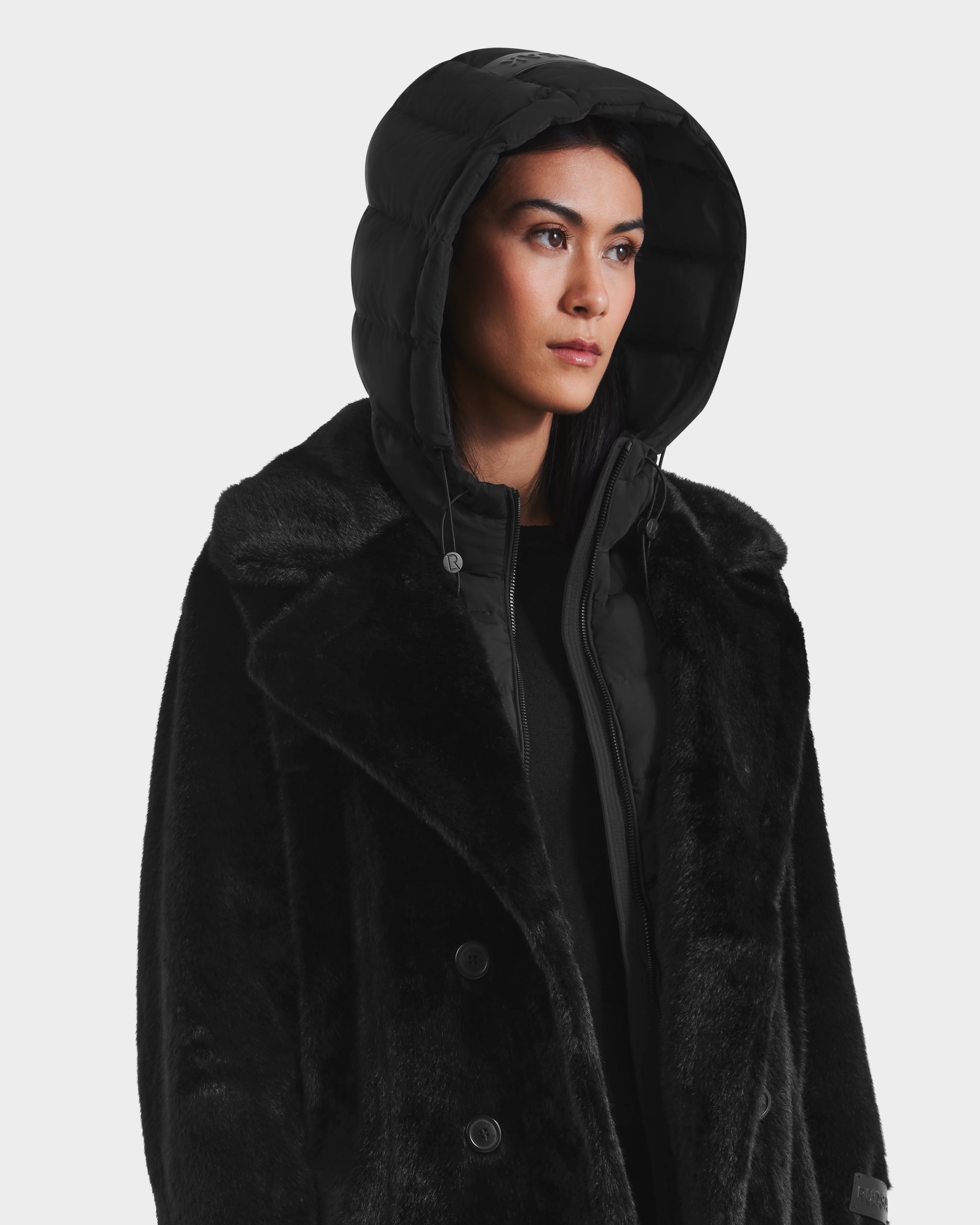 A woman with straight dark hair gazes to the side against a white background, wearing the Rudsak SIENA - 8125542 Black women’s faux fur coat layered over a black down-insulated puffer jacket with the hood up.