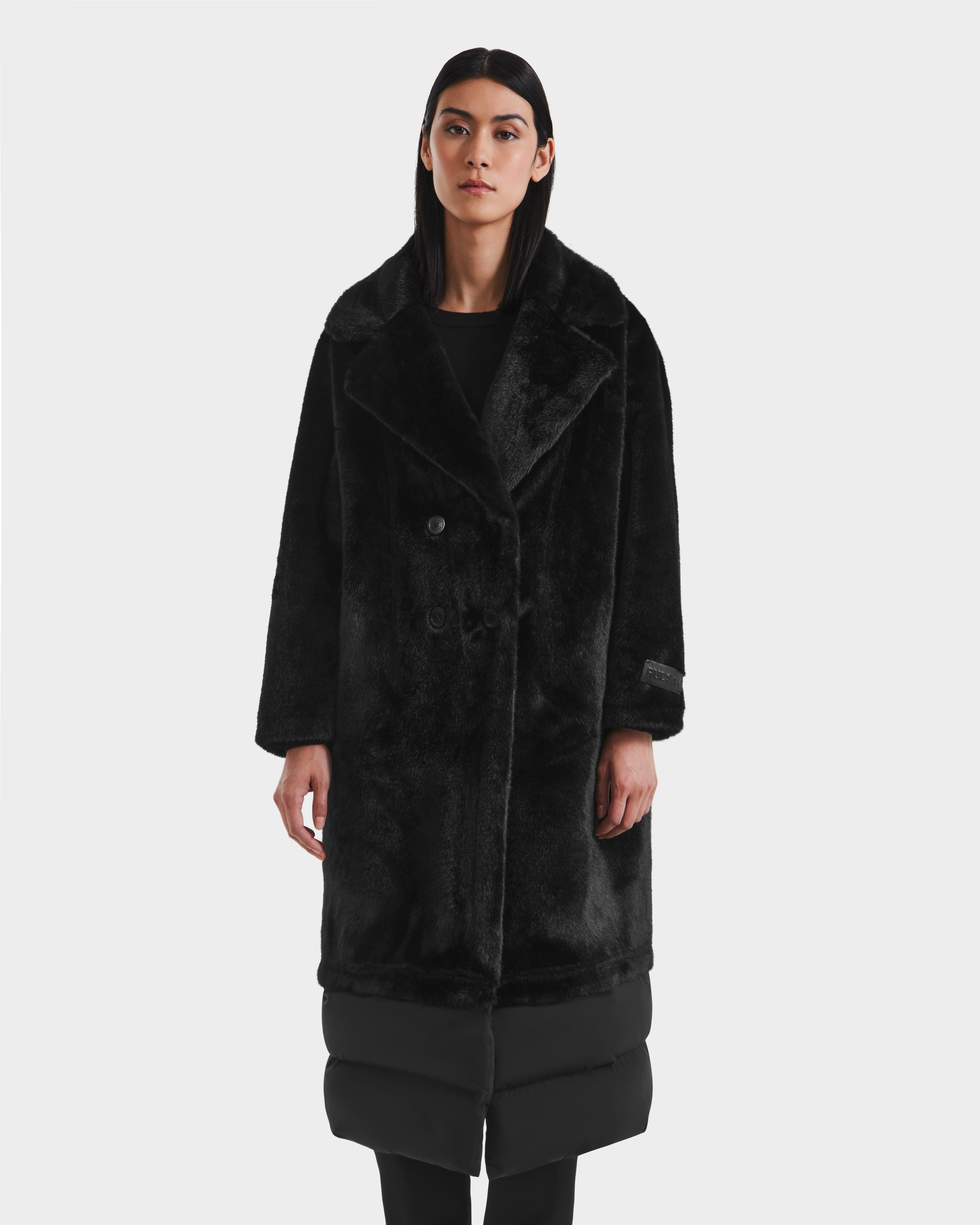 A woman with straight dark hair wears the Rudsak SIENA - 8125542 Black women’s faux fur coat with padded hem, standing against a plain white background—an elegant choice for cold winter days.