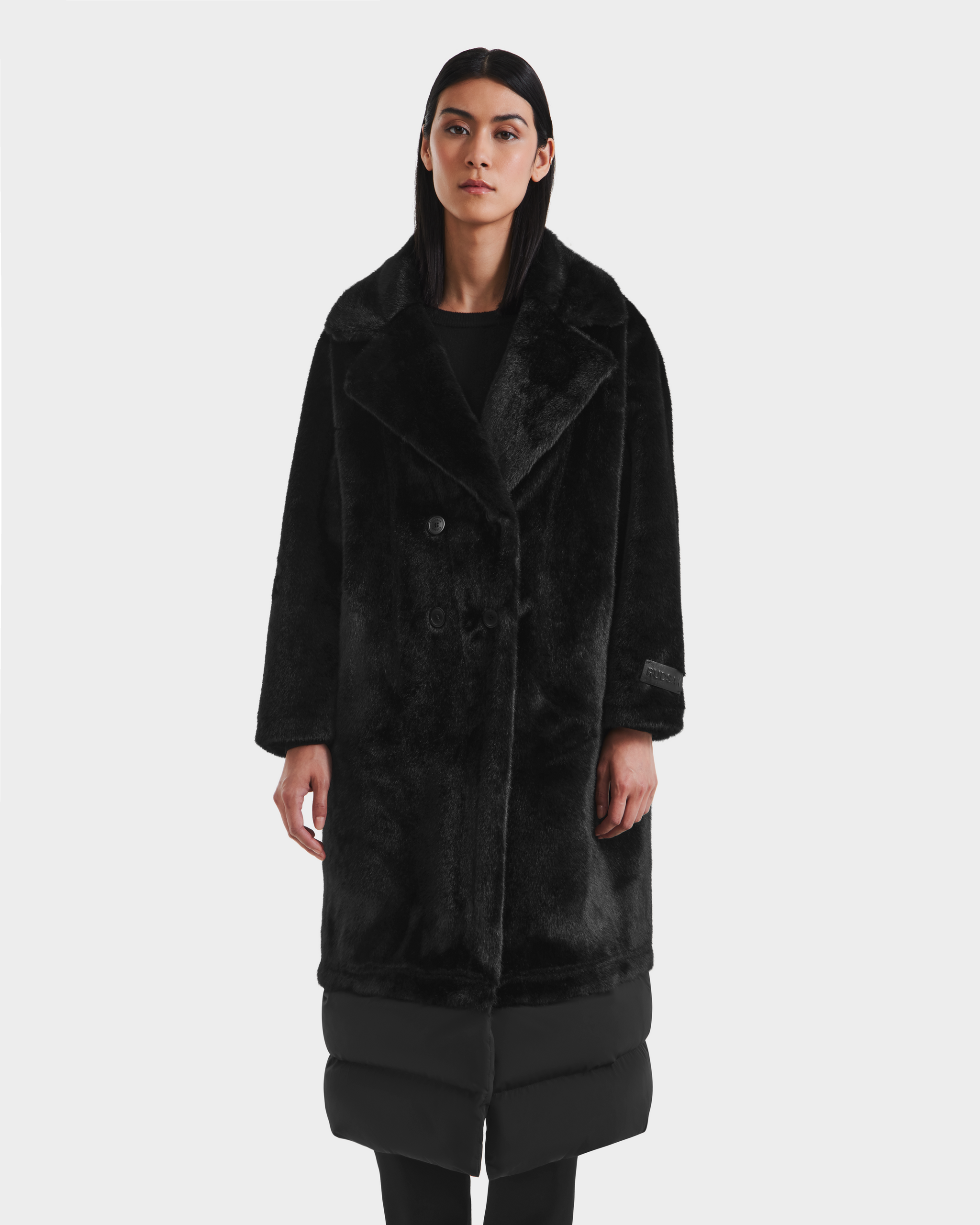 A woman with straight dark hair wears the Rudsak SIENA - 8125542 Black women’s faux fur coat with padded hem, standing against a plain white background—an elegant choice for cold winter days.