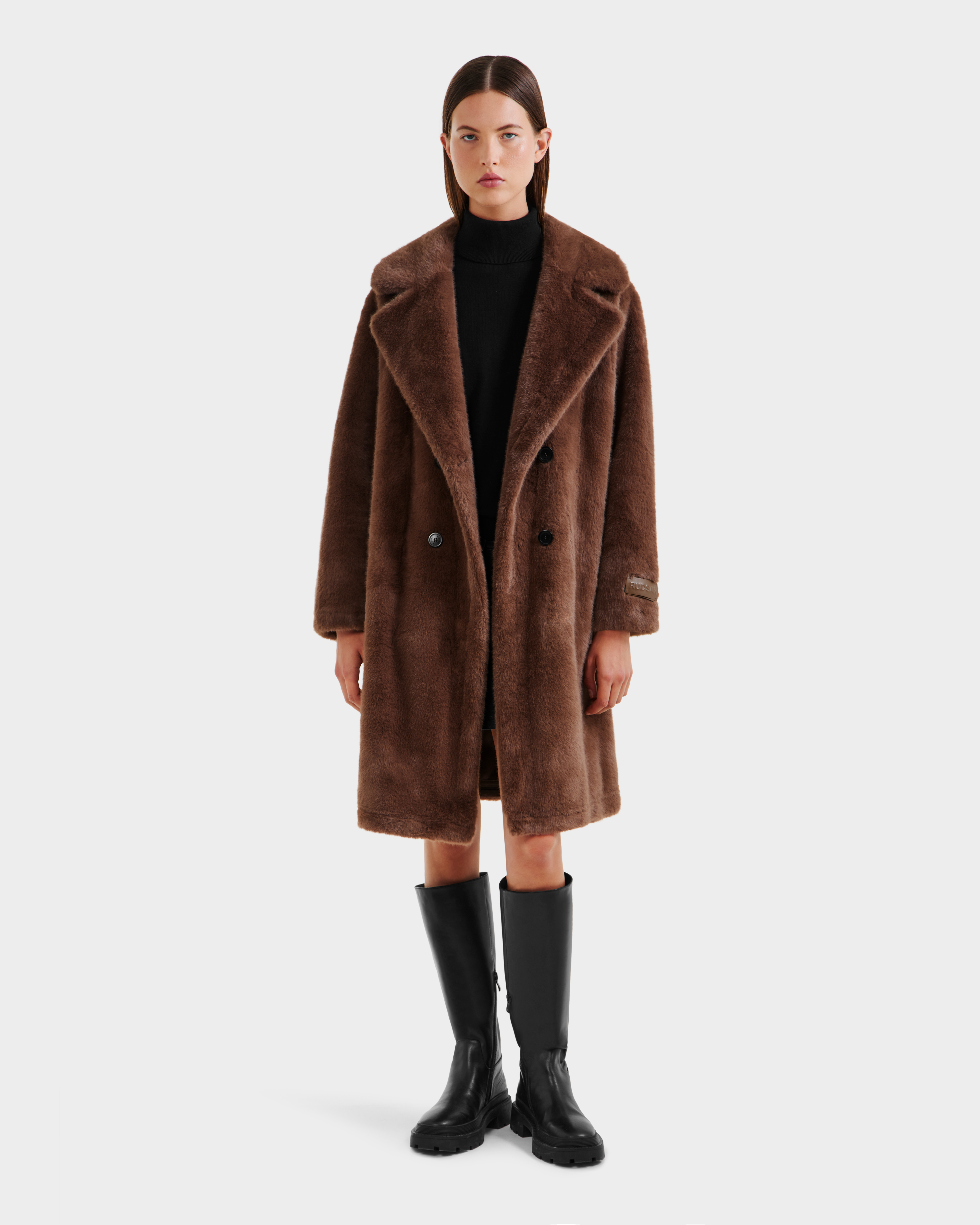 A woman faces forward, wearing the Rudsak SIENA - 8125542 Mocha women's faux fur coat over a black outfit and tall black boots, set against a plain white background.