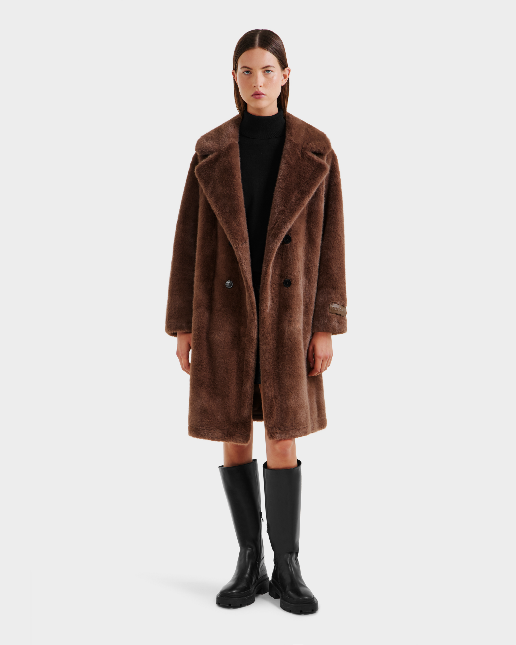 A woman faces forward, wearing the Rudsak SIENA - 8125542 Mocha women's faux fur coat over a black outfit and tall black boots, set against a plain white background.
