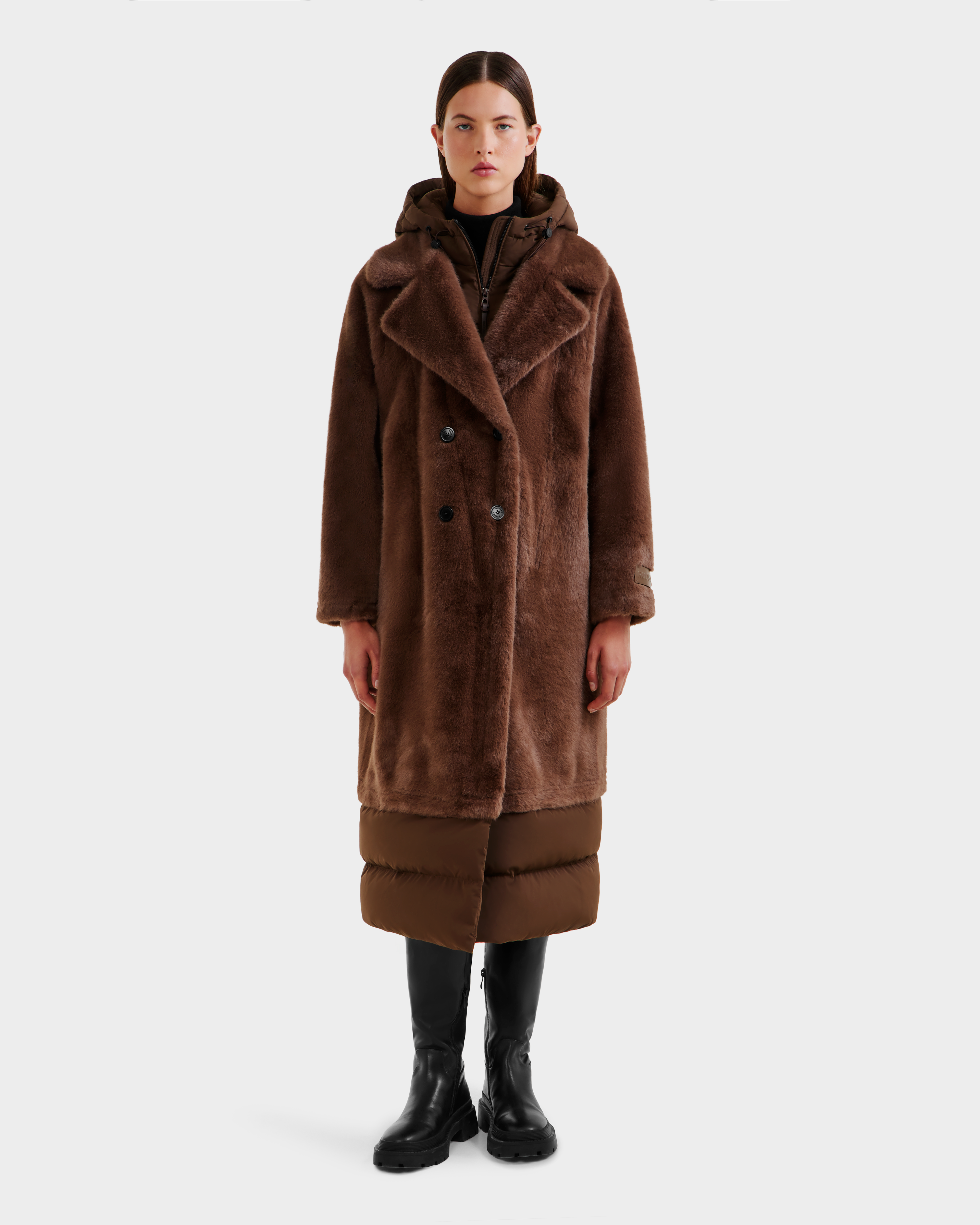 A person stands facing forward against a white background, wearing the Rudsak SIENA - 8125542 Mocha women’s faux fur coat over a puffer jacket, with black knee-high boots and black pants.
