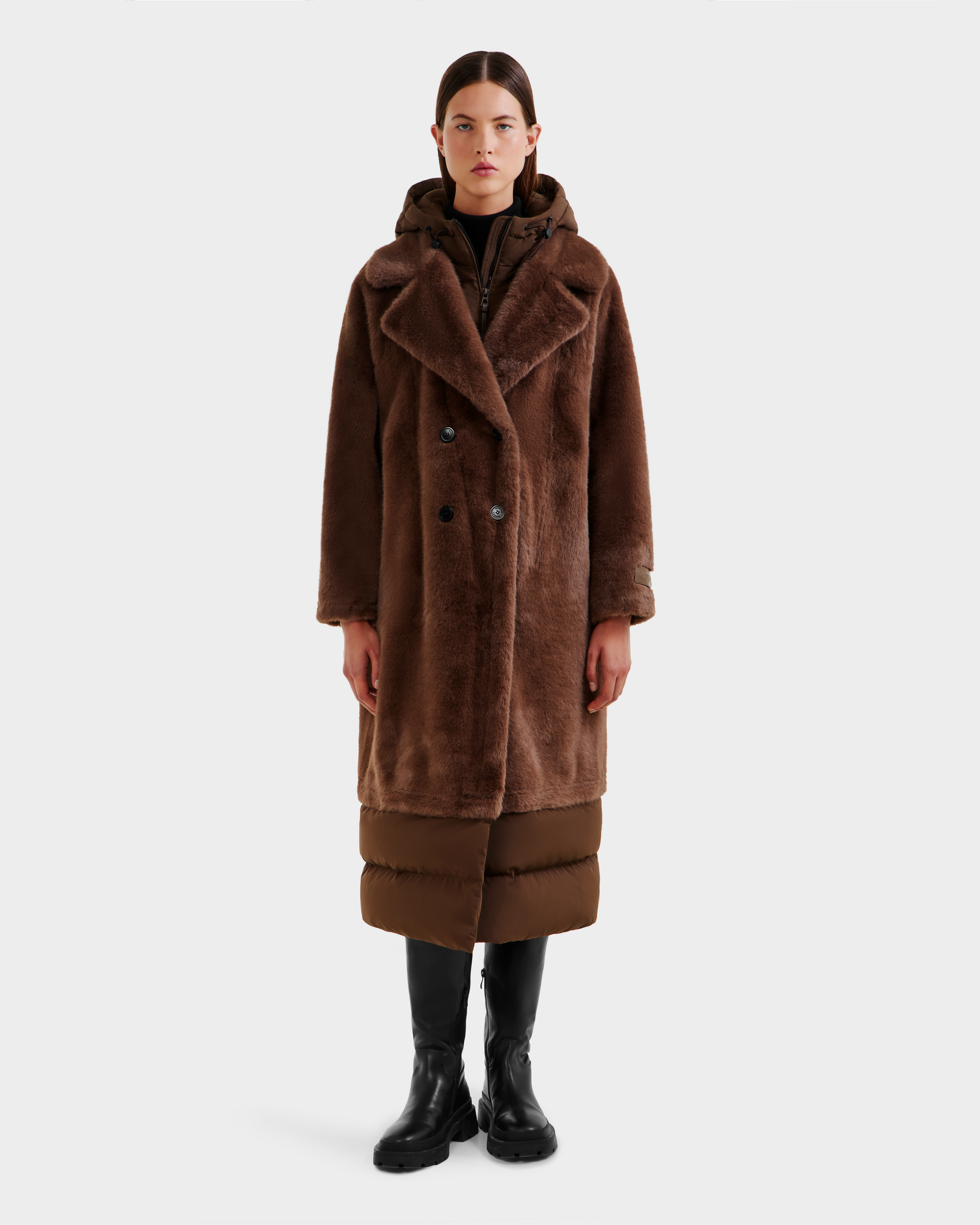 A person stands facing forward against a white background, wearing the Rudsak SIENA - 8125542 Mocha women’s faux fur coat over a puffer jacket, with black knee-high boots and black pants.