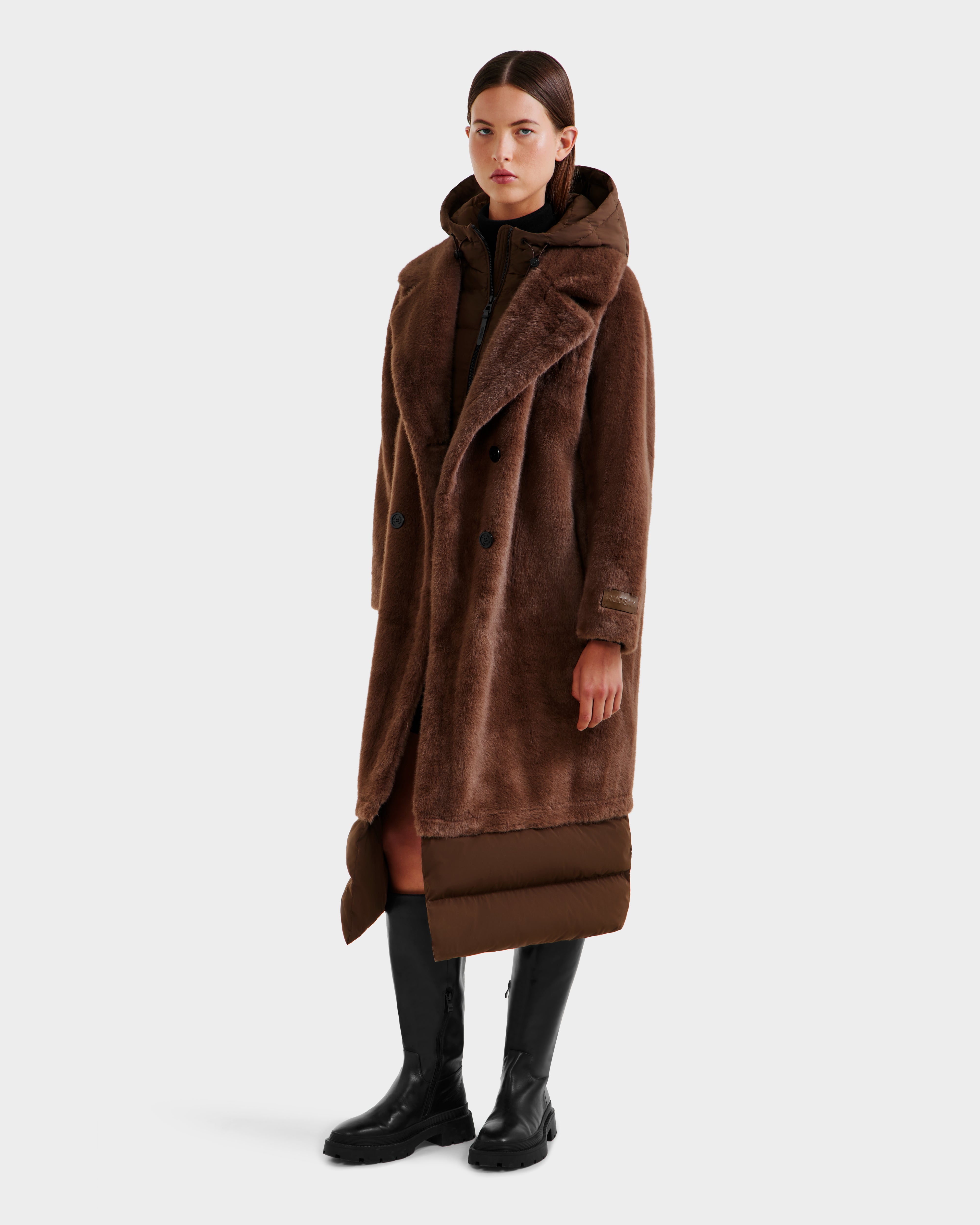 A woman stands against a plain background wearing the Rudsak SIENA - 8125542 Mocha women’s faux fur coat layered over a down-insulated coat with removable hooded dickie, paired with black knee-high boots and a neutral expression.