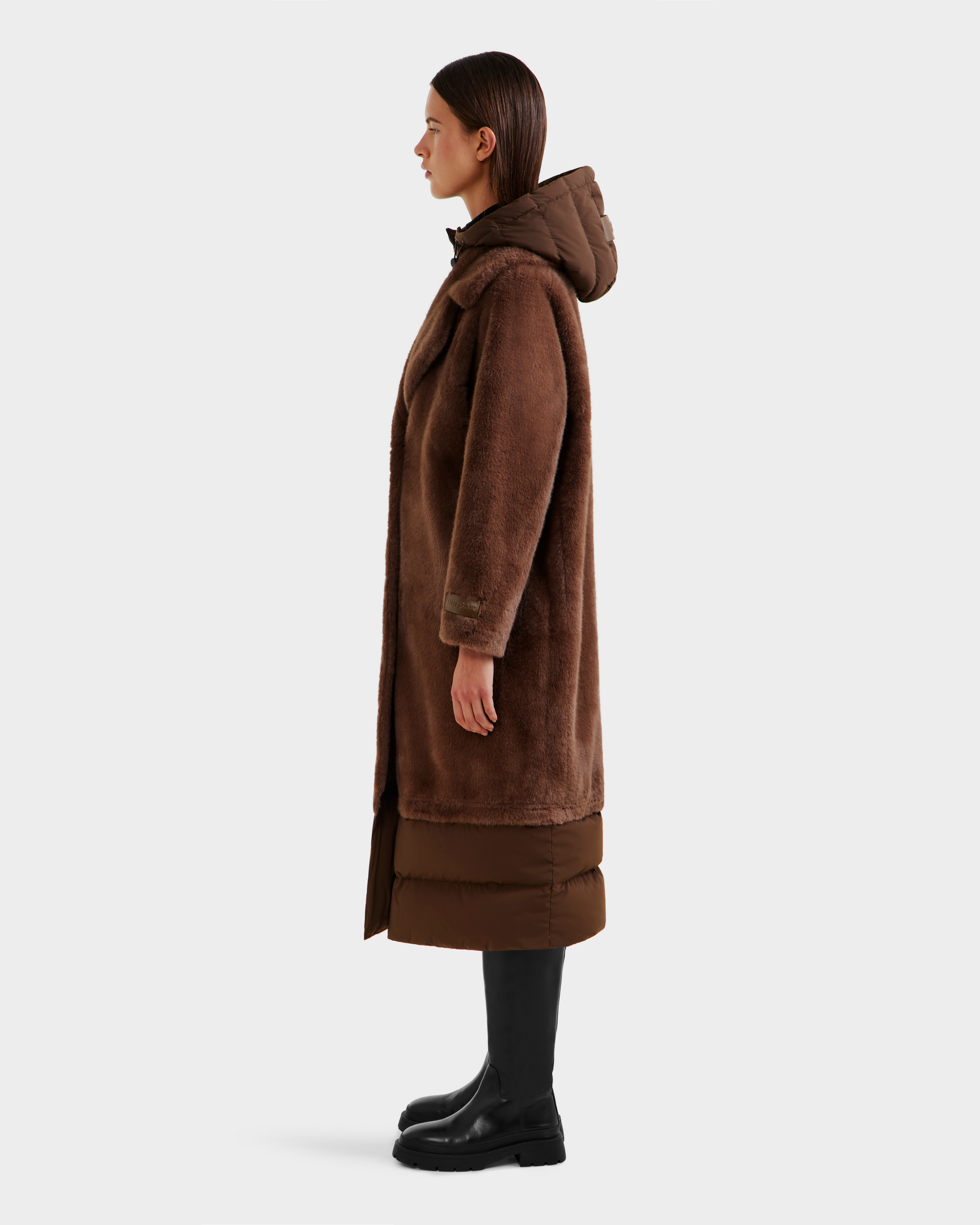 A woman in profile wears the Rudsak SIENA - 8125542 Mocha women’s faux fur coat with a hood and brown quilted hem, paired with black knee-high boots and dark pants, standing against a plain light background.
