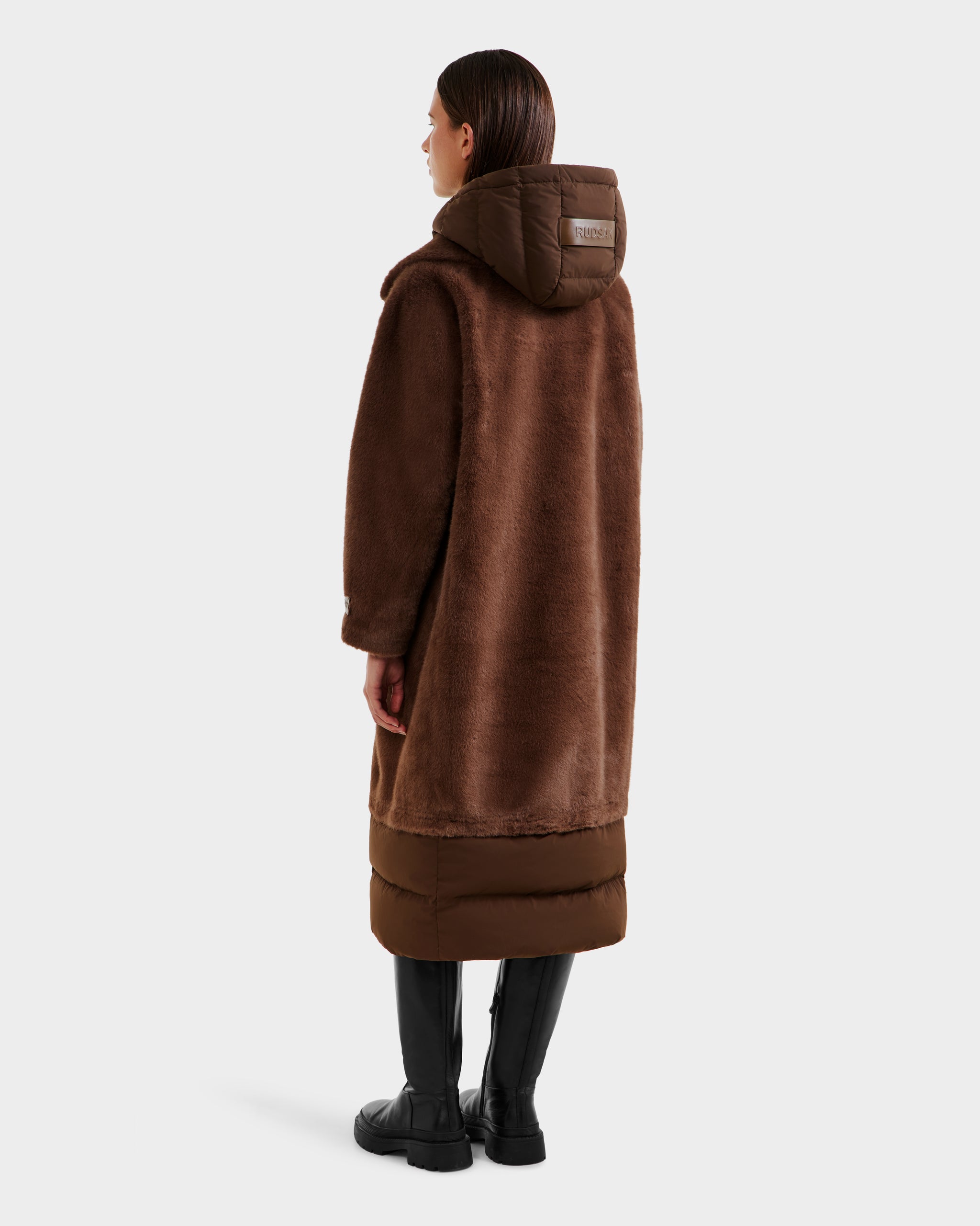 A person stands facing away, wearing the Rudsak SIENA - 8125542 Mocha womens faux fur coat with a puffer-style hood and lower section, paired with black knee-high boots against a plain light background.