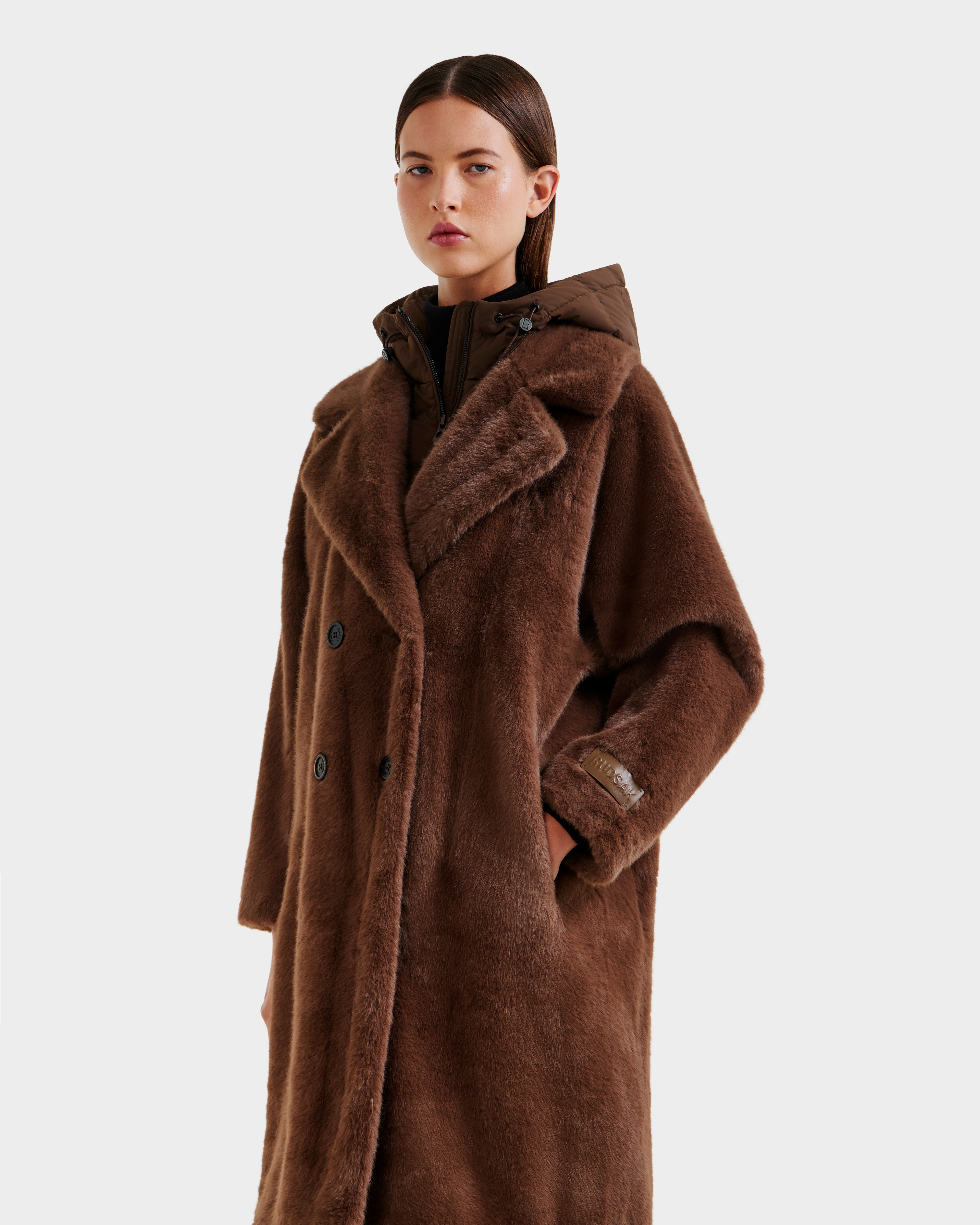 A woman with straight brown hair models the Rudsak SIENA - 8125542 Mocha women’s faux fur coat, featuring a wide collar and button details, layered over a down-insulated hooded dickie, against a plain white background.