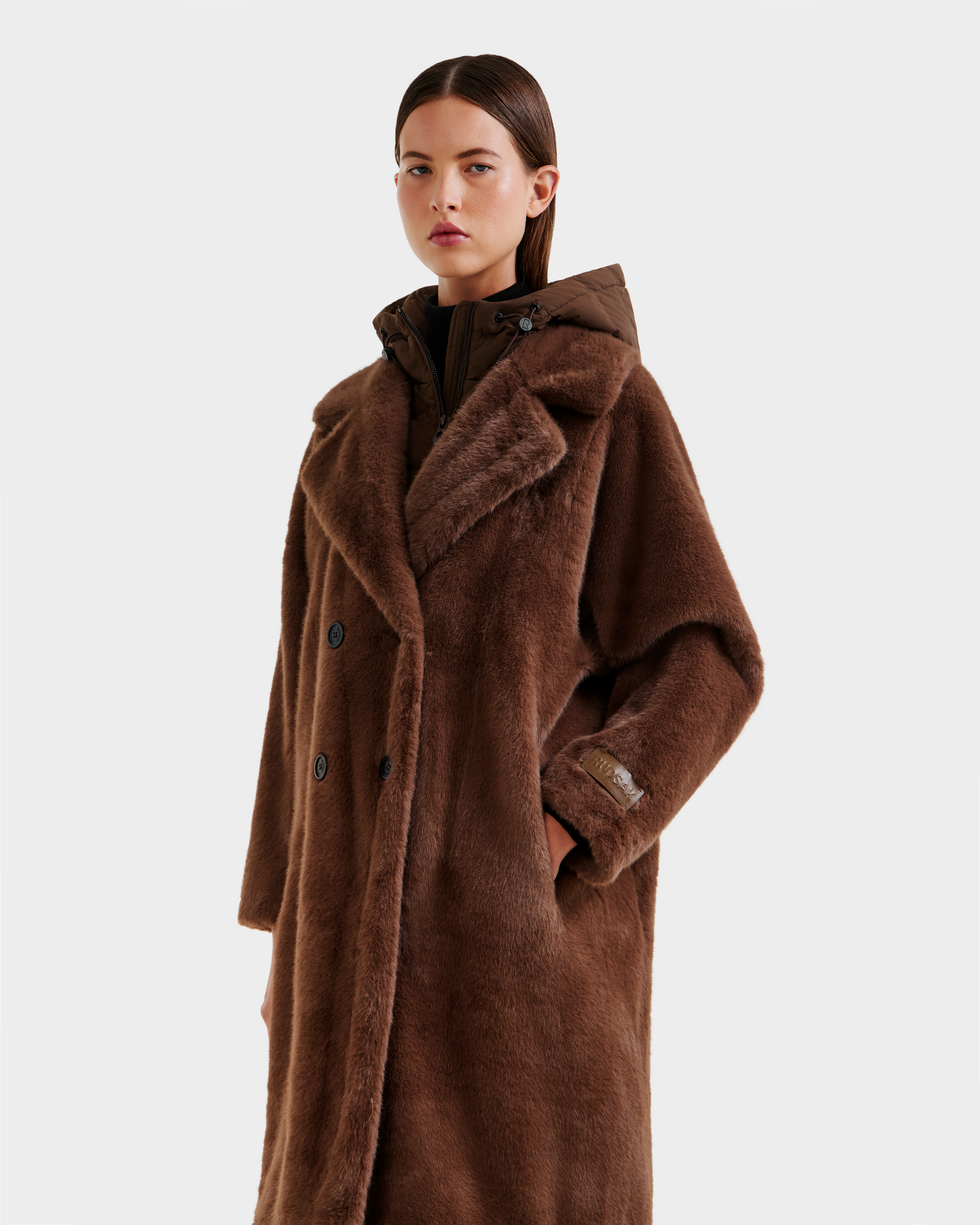 A woman with straight brown hair models the Rudsak SIENA - 8125542 Mocha women’s faux fur coat, featuring a wide collar and button details, layered over a down-insulated hooded dickie, against a plain white background.