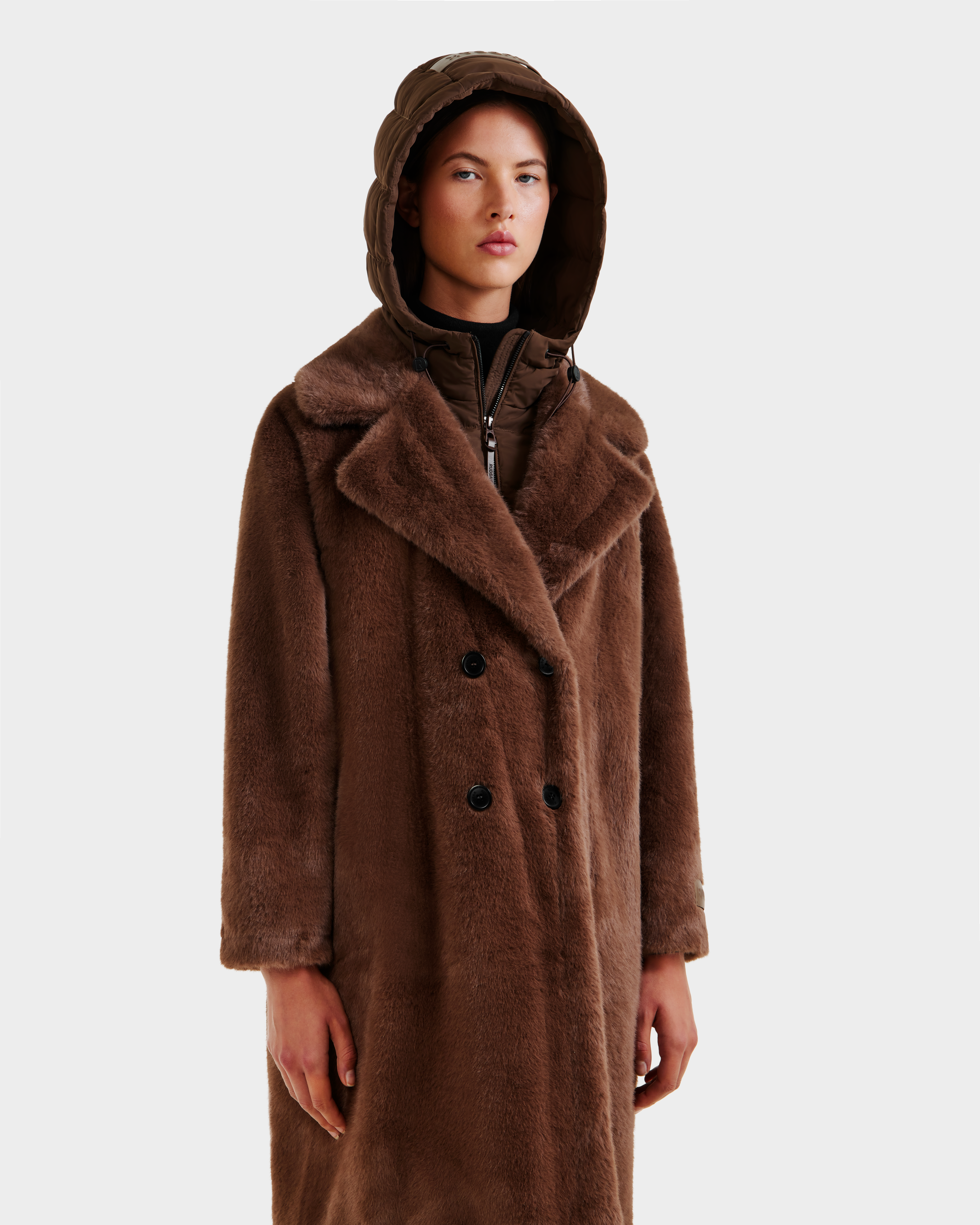A person wears the Rudsak SIENA - 8125542 Mocha women's faux fur coat with wide lapels and a quilted hooded layer underneath, standing against a plain light background.