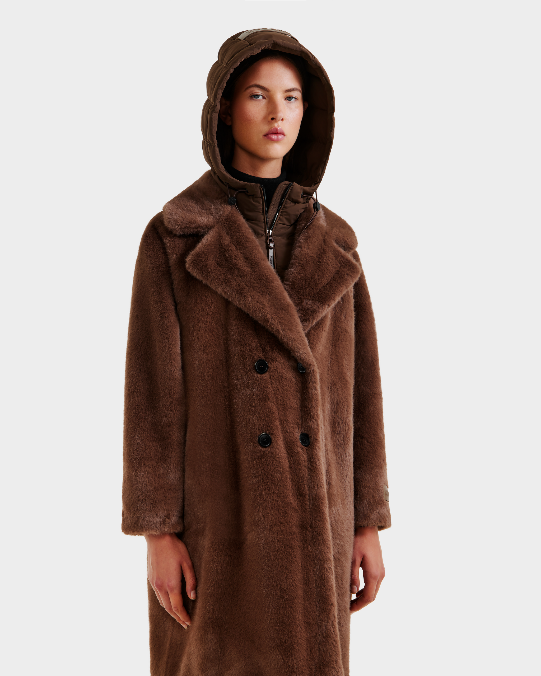 A person wears the Rudsak SIENA - 8125542 Mocha women's faux fur coat with wide lapels and a quilted hooded layer underneath, standing against a plain light background.