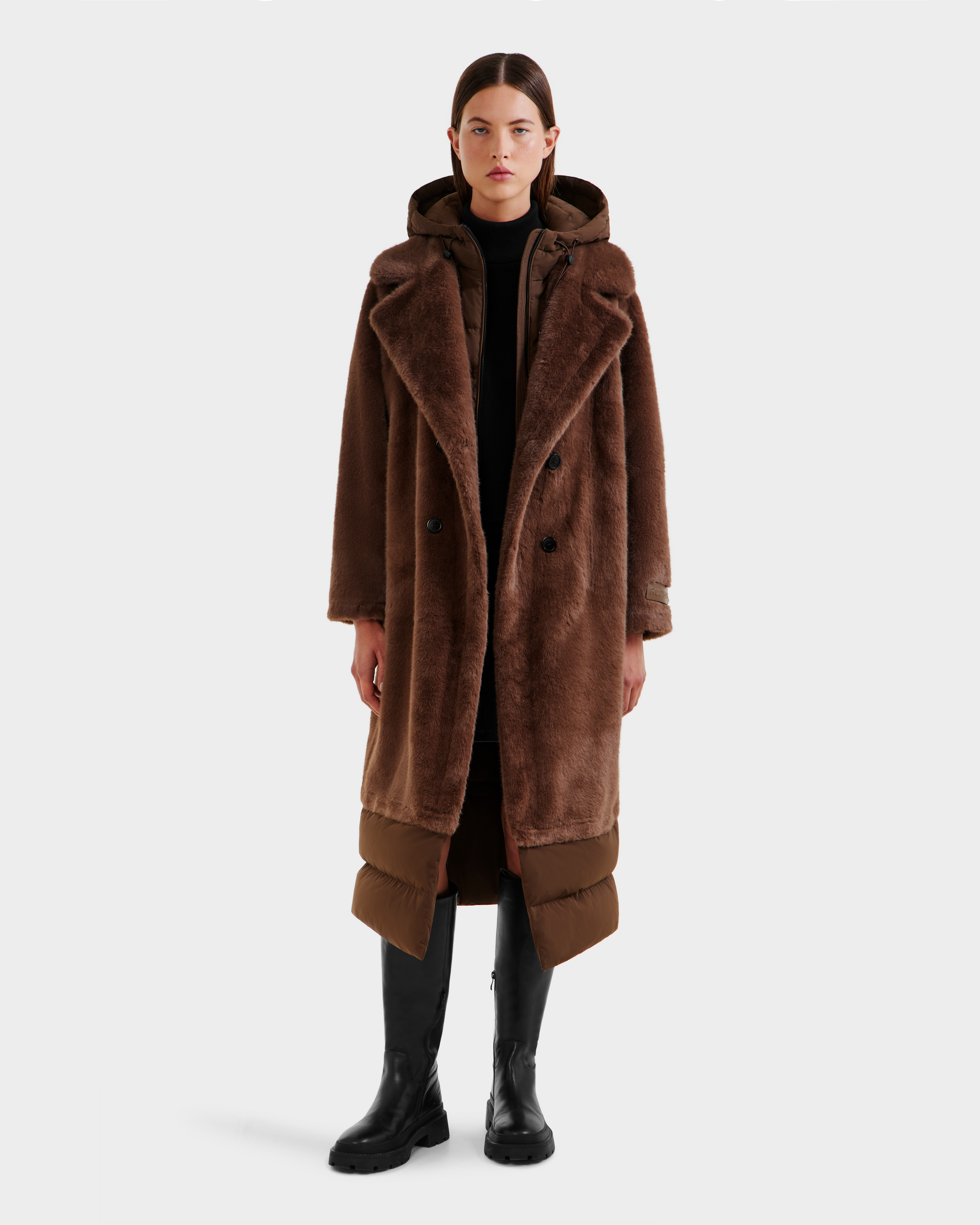 A woman faces forward wearing the Rudsak SIENA - 8125542 Mocha women’s faux fur coat with oversized lapels, hood, black boots, and a dark turtleneck dress. The plain white background highlights the chic winter style.